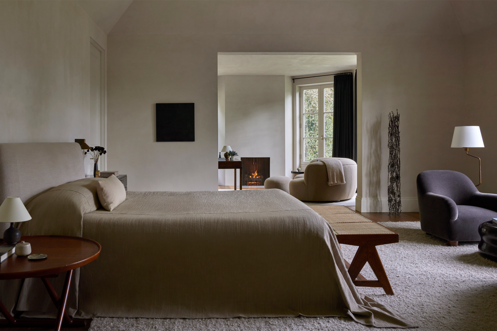 A cozy bedroom with a large bed, nightstand with a lamp and decorative items, and a sitting area with a plush chair, small table, and a floor lamp, with natural light coming through tall windows.