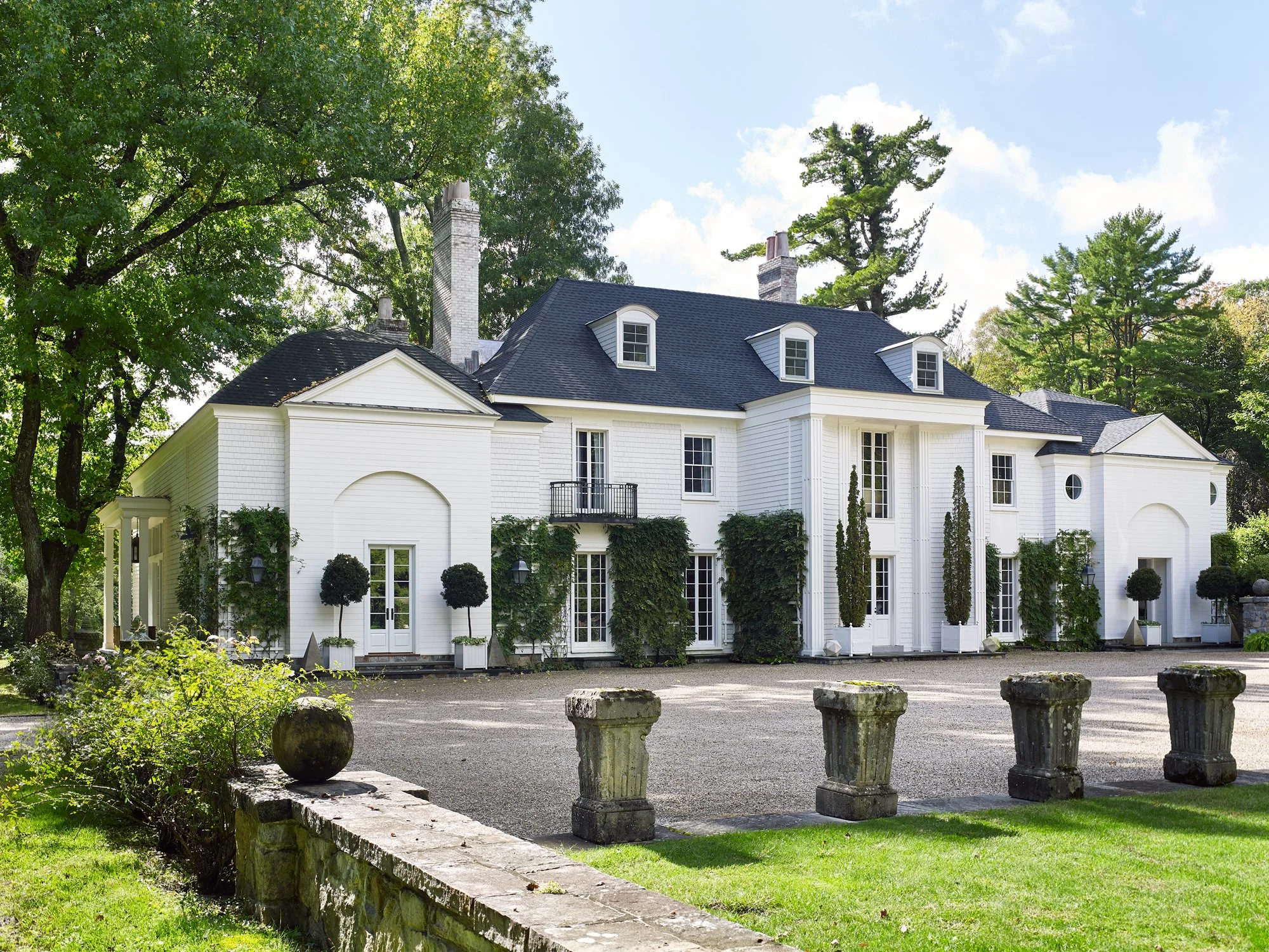 A large, white, two-story house with a dark roof, multiple windows, French doors, and a small balcony, surrounded by tall trees and a landscaped garden with potted plants and ivy, in a lush, green setting.