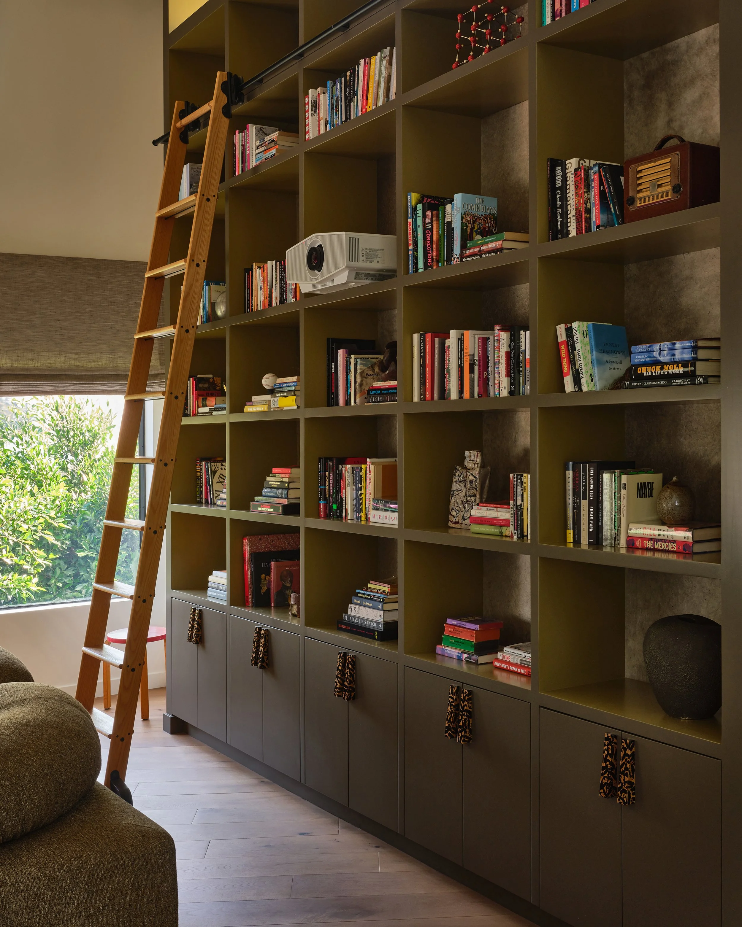 Comedian Anthony Jeselnik’s library, interior design by Riccio Vidaurre Design – A large olive green bookshelf with a rolling library ladder and hardware pulls by atelier Jonathan cohen.
