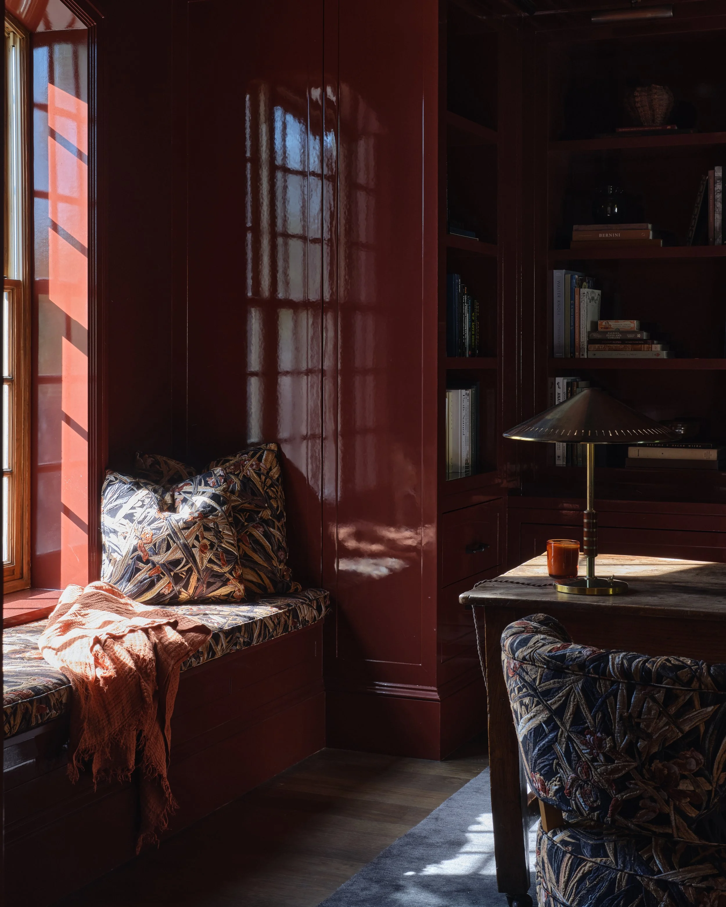 A cozy interior corner with a window seat, patterned cushions, a blanket, a desk lamp, a wooden table, a cup, and a bookshelf filled with books in a room with dark wood walls and flooring.