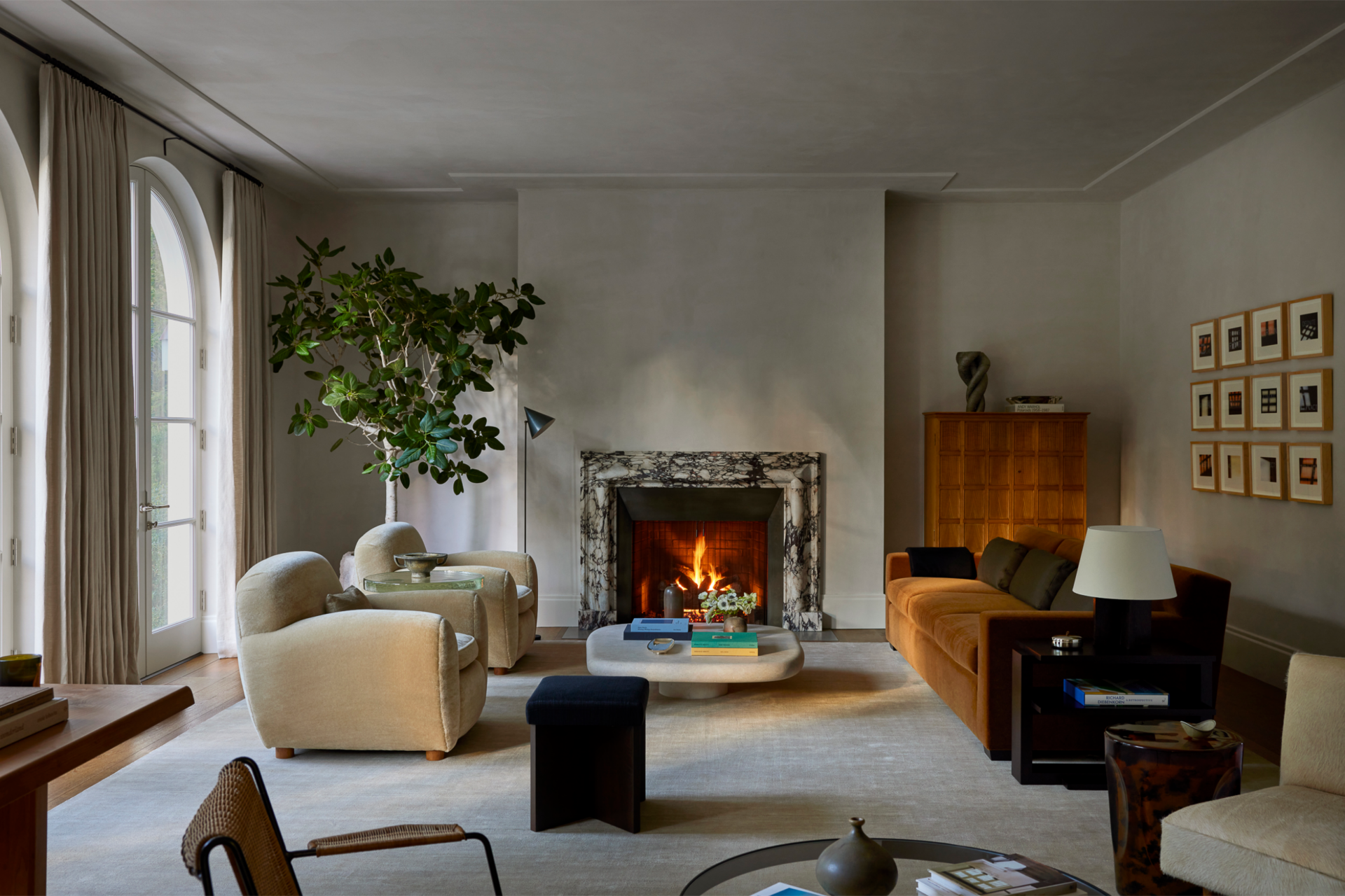 Living room with a fireplace, beige chairs, a large plant, and artwork on the wall.