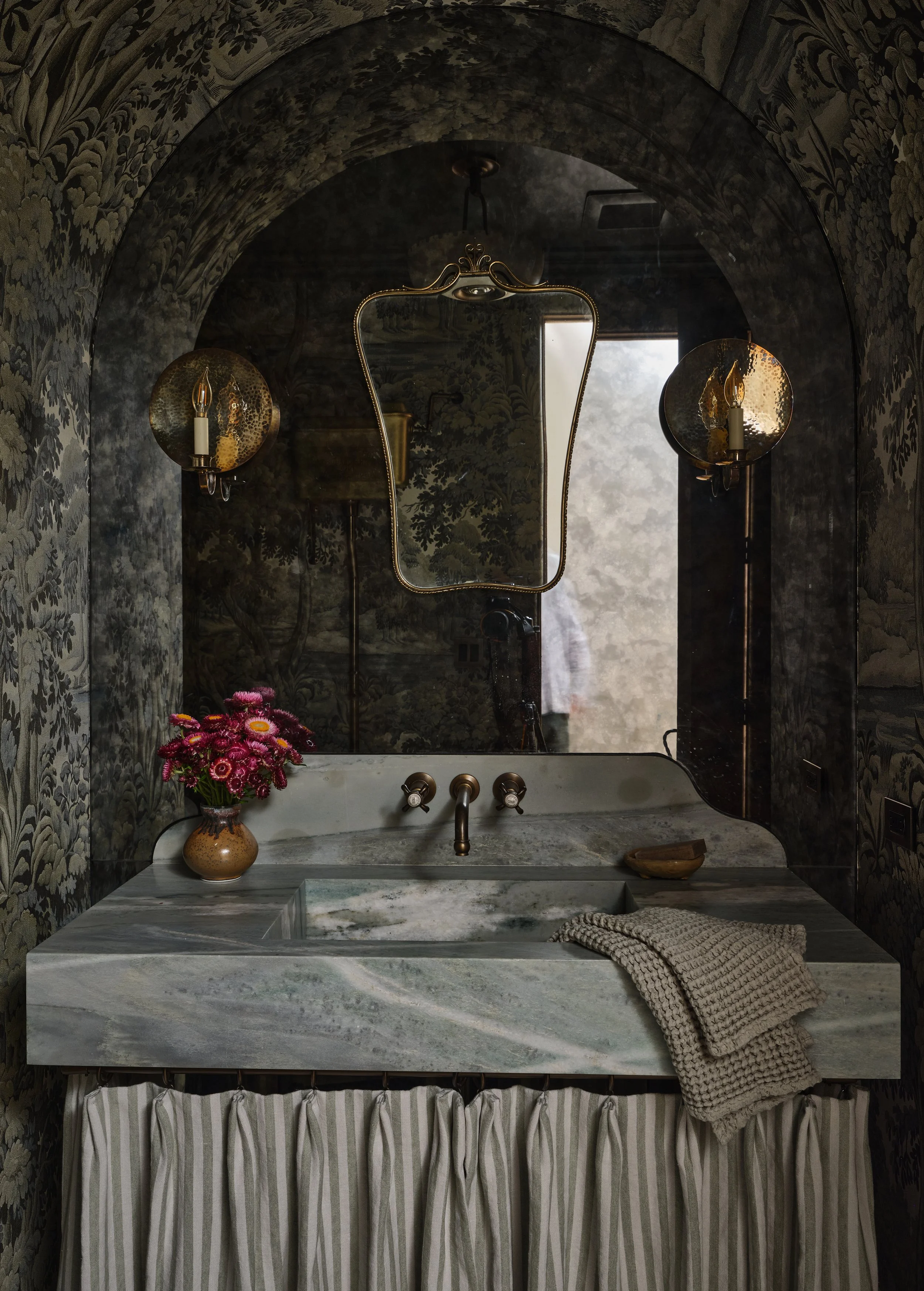 A vintage bathroom with a marble sink, floral wallpaper, a mirror, two wall sconces, and a pink flower arrangement on the counter.