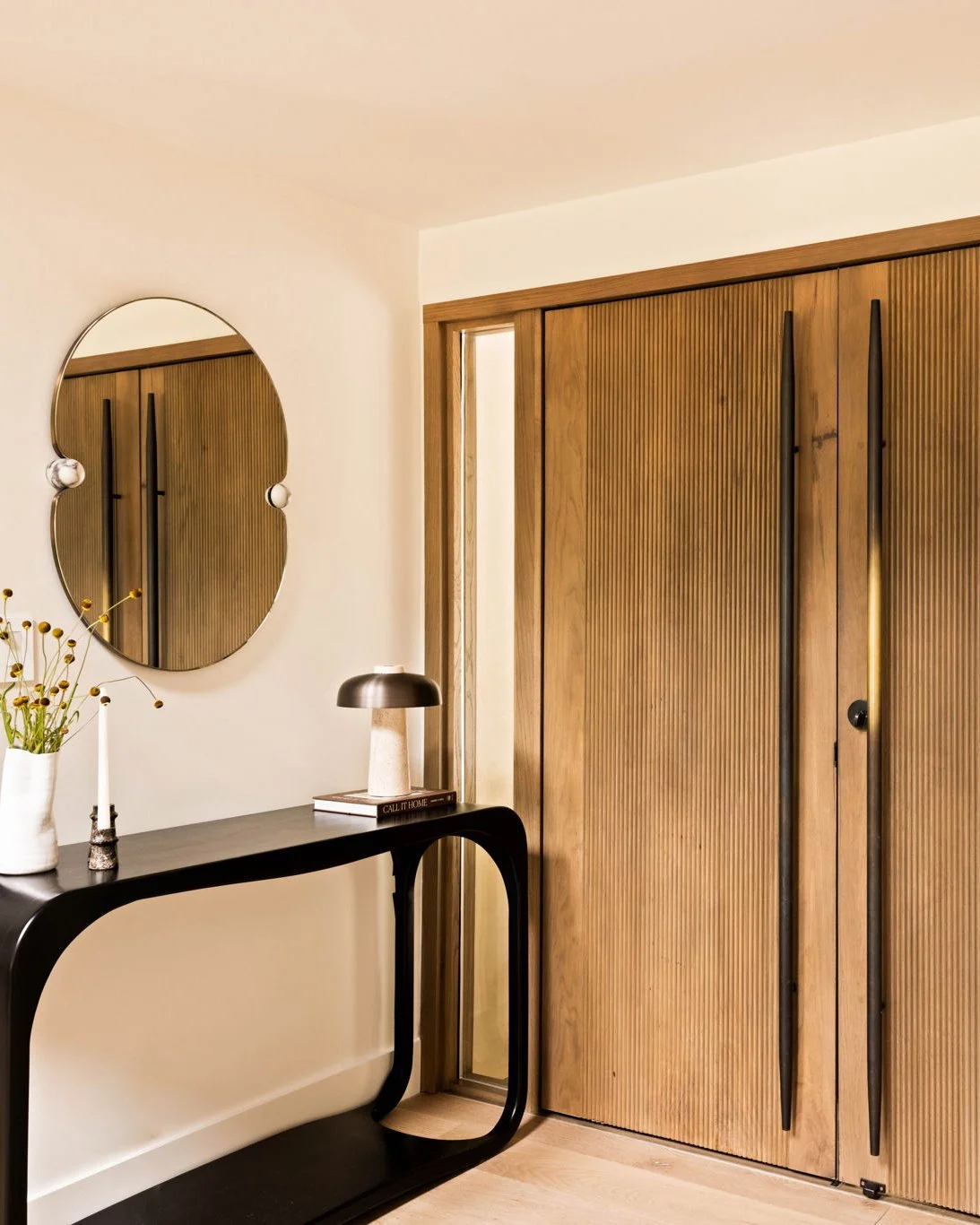 An entryway of a mid-century ranch house with white plaster walls and a ribbed wood entry door with long blackened brass handles. There is a black console table with a table lamp and a vase of flowers with a organic shaped mirror hanging above.