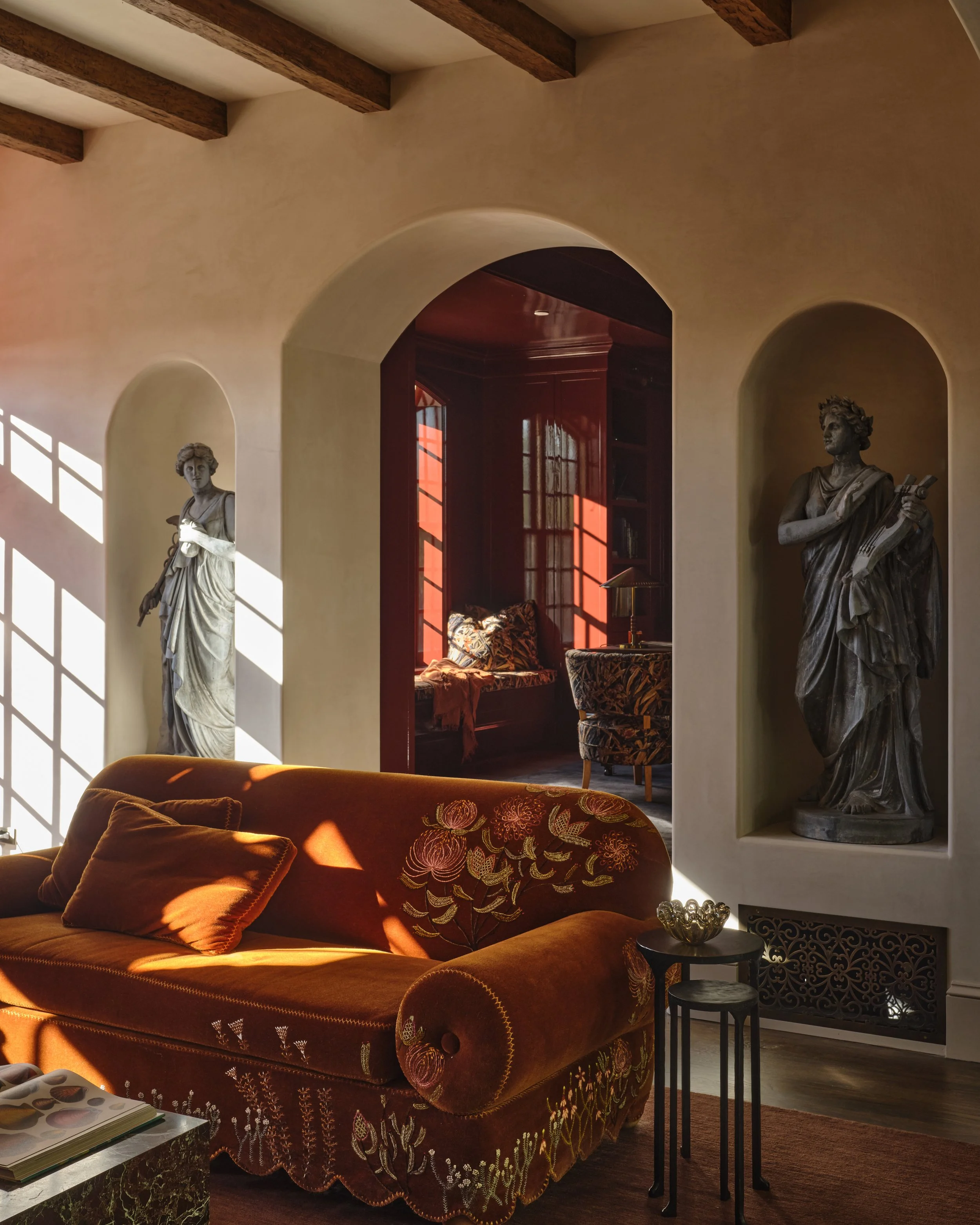 Living room with sunlight casting shadows on the wall, featuring a burnt orange velvet sofa with embroidered floral designs, decorative pillows, two classical statues in wall niches, and a cozy seating area with a window seat and armchair in the background.