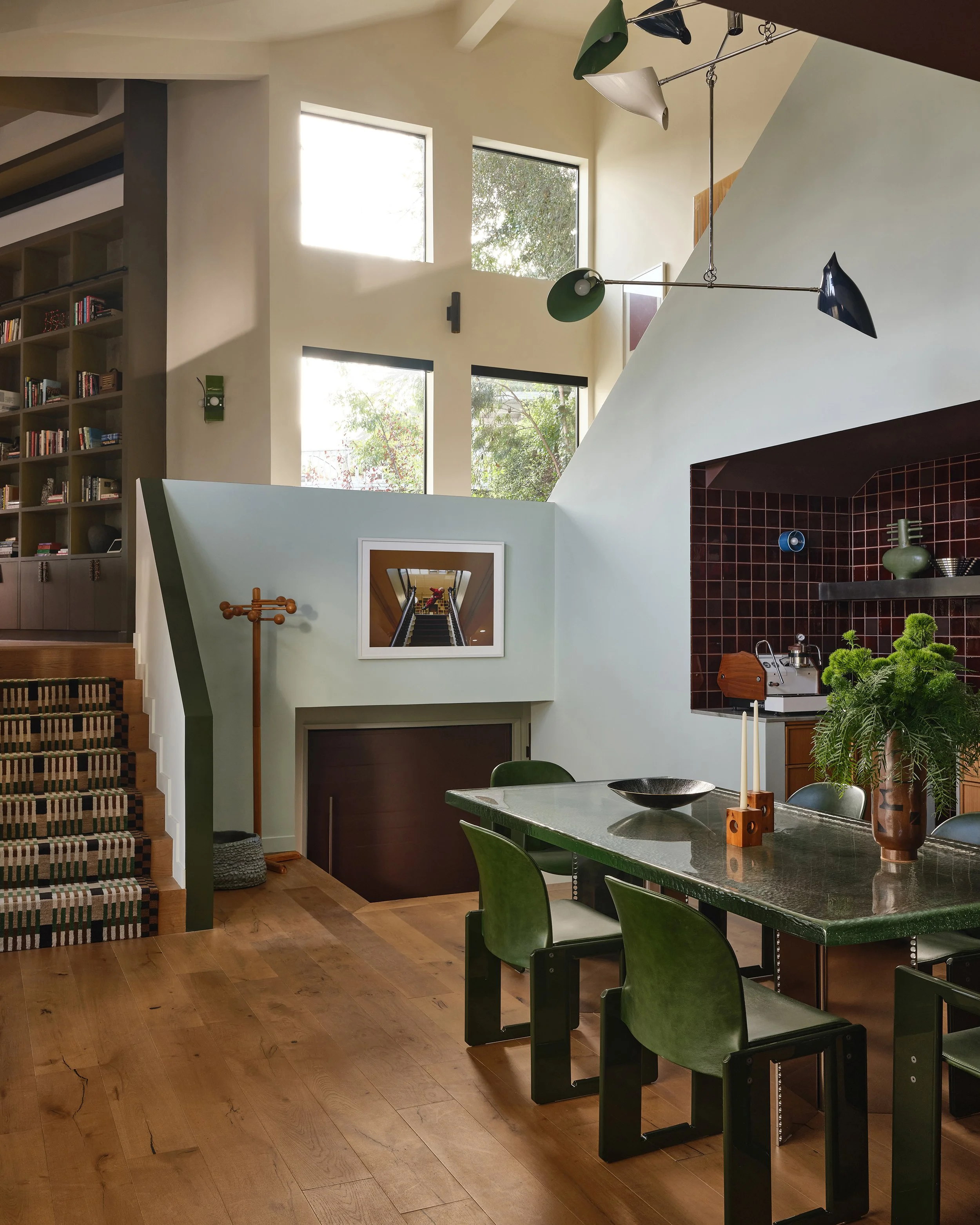 Comedian Anthony Jeselnik’s home by Riccio Vidaurre Design – A eat in kitchen with a cast glass dining table and green dialogo dining chairs by Scarpa.  Light blue walls, green handrail and a geometric patterned stair runner.