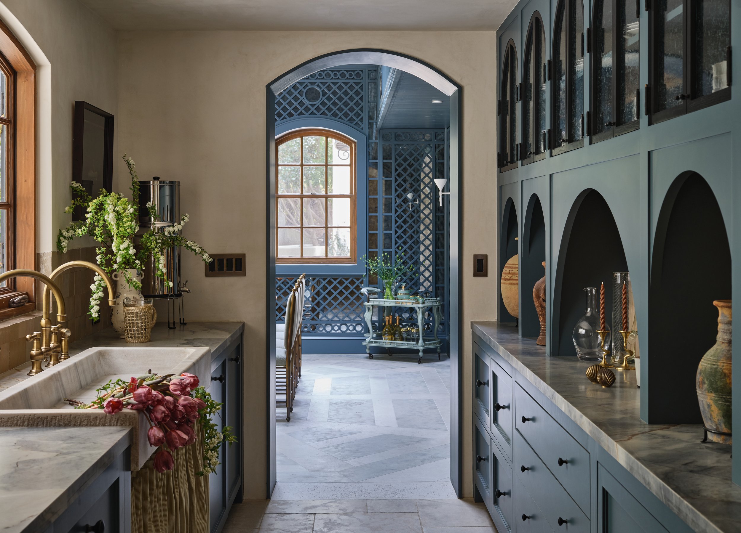 Interior view of a kitchen with a marble countertop, gold sink fixtures, and glass cabinets. Decor includes vases, candles, and flowers. A doorway leads to a sunlit room with blue walls and a window, decorated with lattice details.