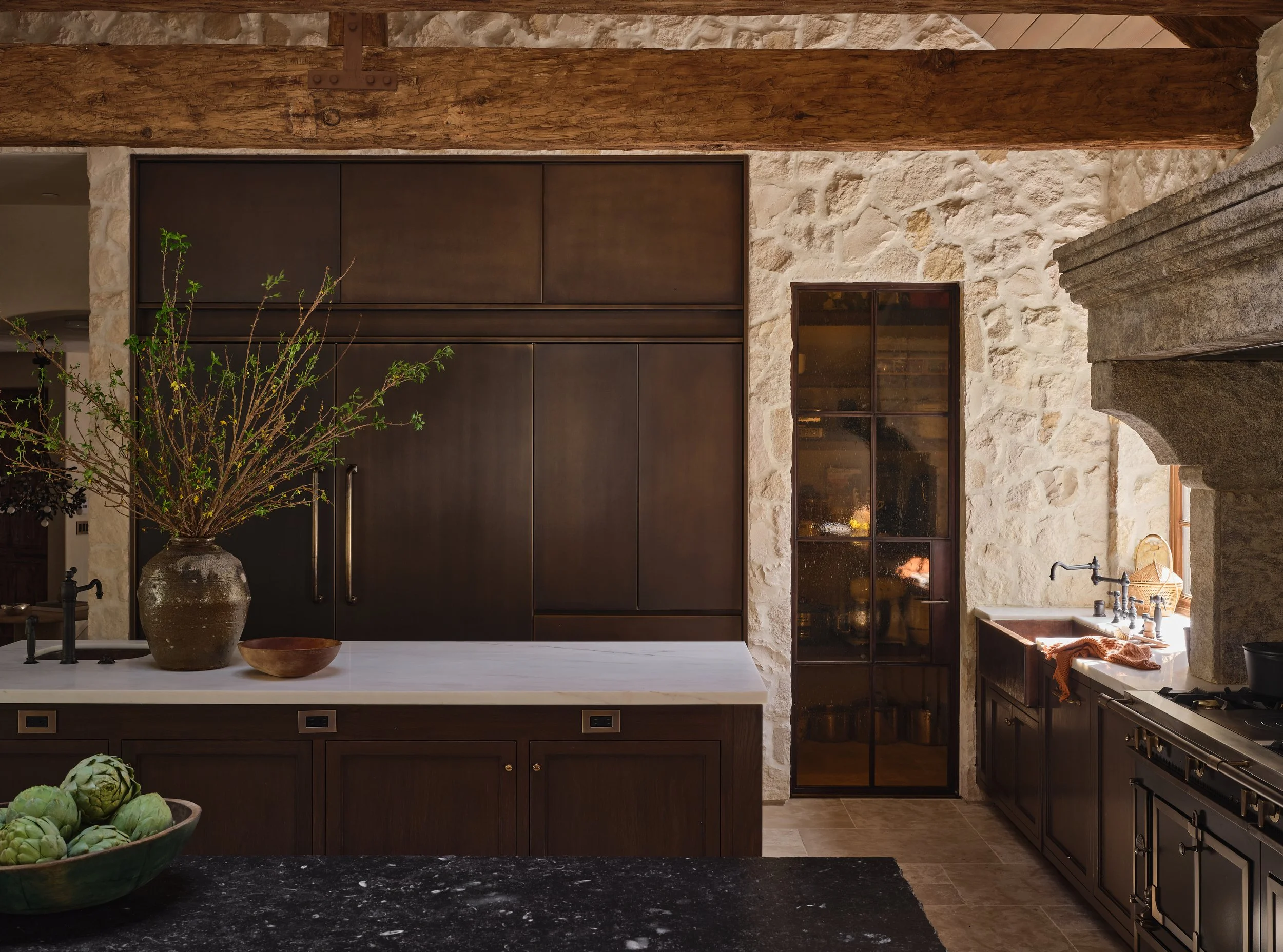 A rustic kitchen with stone walls, wooden beams, dark wood cabinets, a white marble countertop, a large vase with branches, a bowl of artichokes, and a window with a view into an adjacent room.