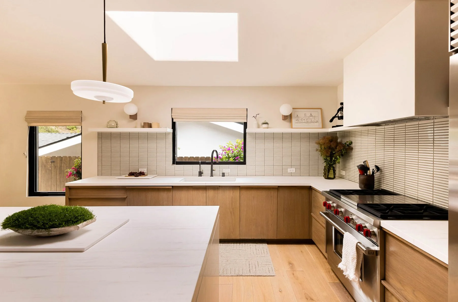 A remodeled kitchen in a mid-century modern home.  Walnut cabinets with stacked white finger tiles on the backsplash.  A white shelf above holds glasses and art.  There is a plastered hood above the range and a large marble island.