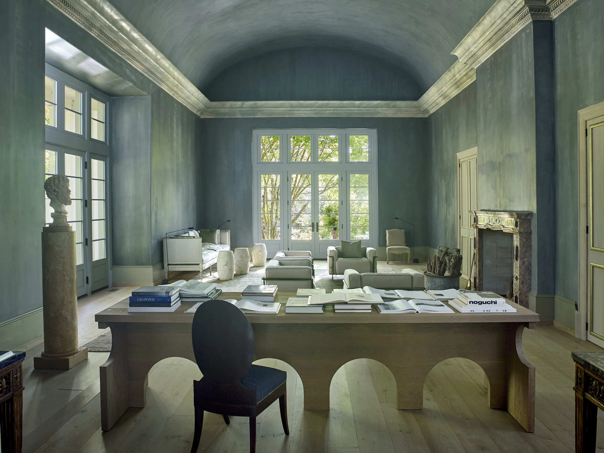 Bright living room with high, arched ceiling, large windows, and a mix of modern and classical decor. A wooden desk with open books and folders is in the foreground, with a black chair. The room has white sofas, stone bust sculpture on a pedestal, and a fireplace.