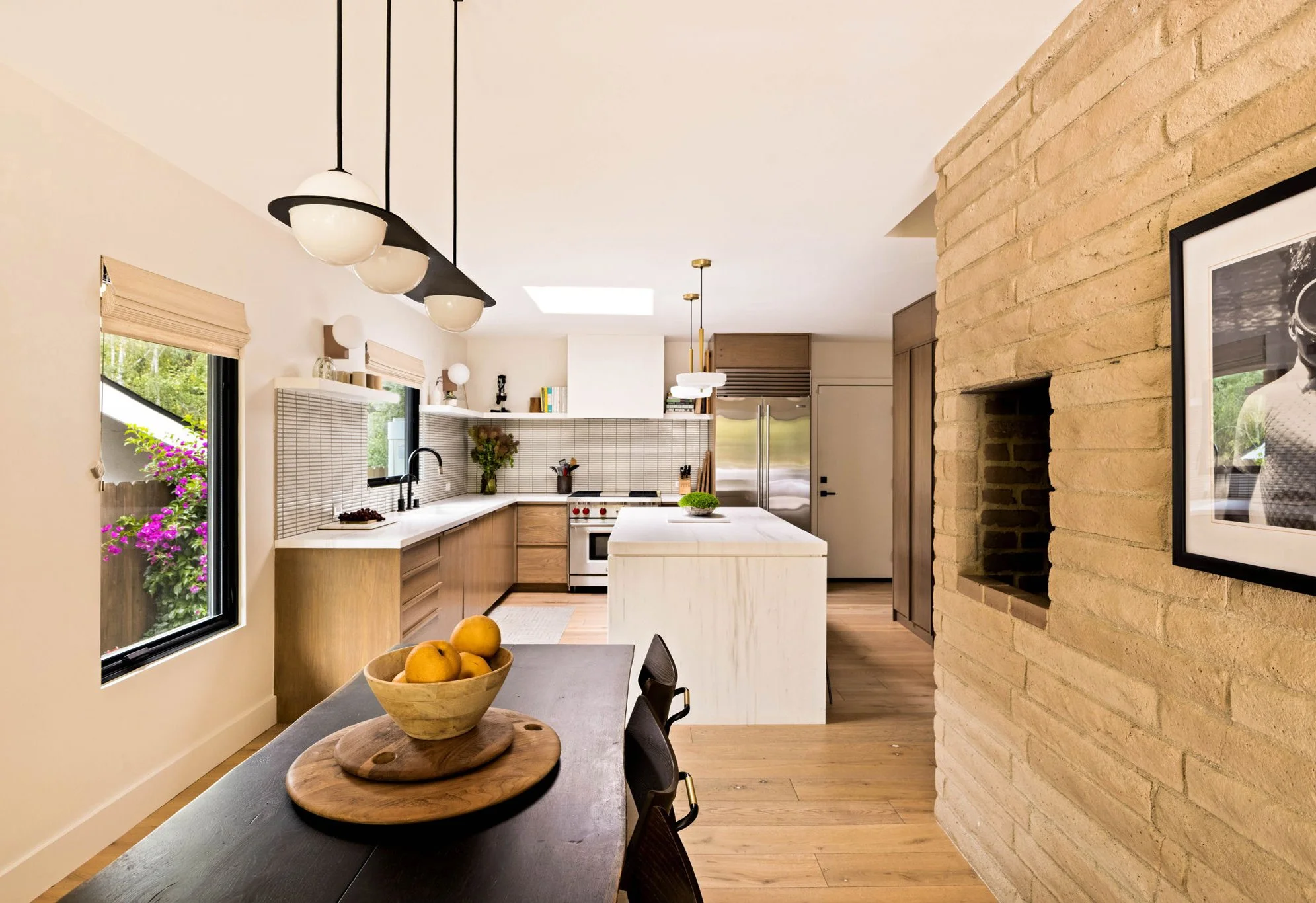 A dining room and kitchen in a mid-century modern home.  The dining room has a cream brick wall, black wood dining table and a linear pendant.  The kitchen has been redesigned and is modern with white stone, white tile and walnut cabinetry.