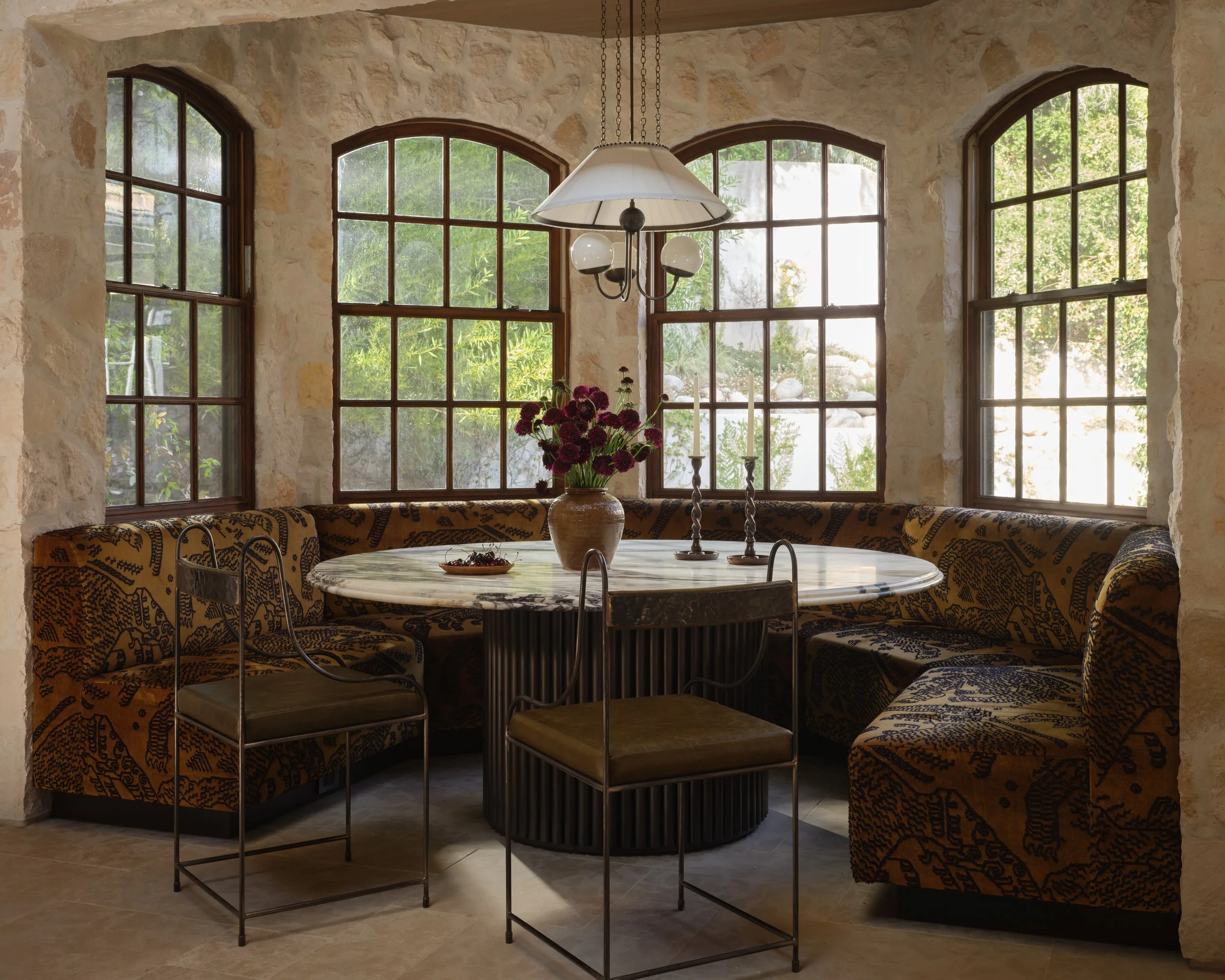 A dining nook with a round marble table, surrounded by upholstered banquette seating with a black and gold pattern. The table has a large ceramic vase with purple flowers, two tall candlesticks, and a hanging vintage-style pendant light. Behind the table are large, arched windows with wooden frames, letting in natural light and views of greenery outside.