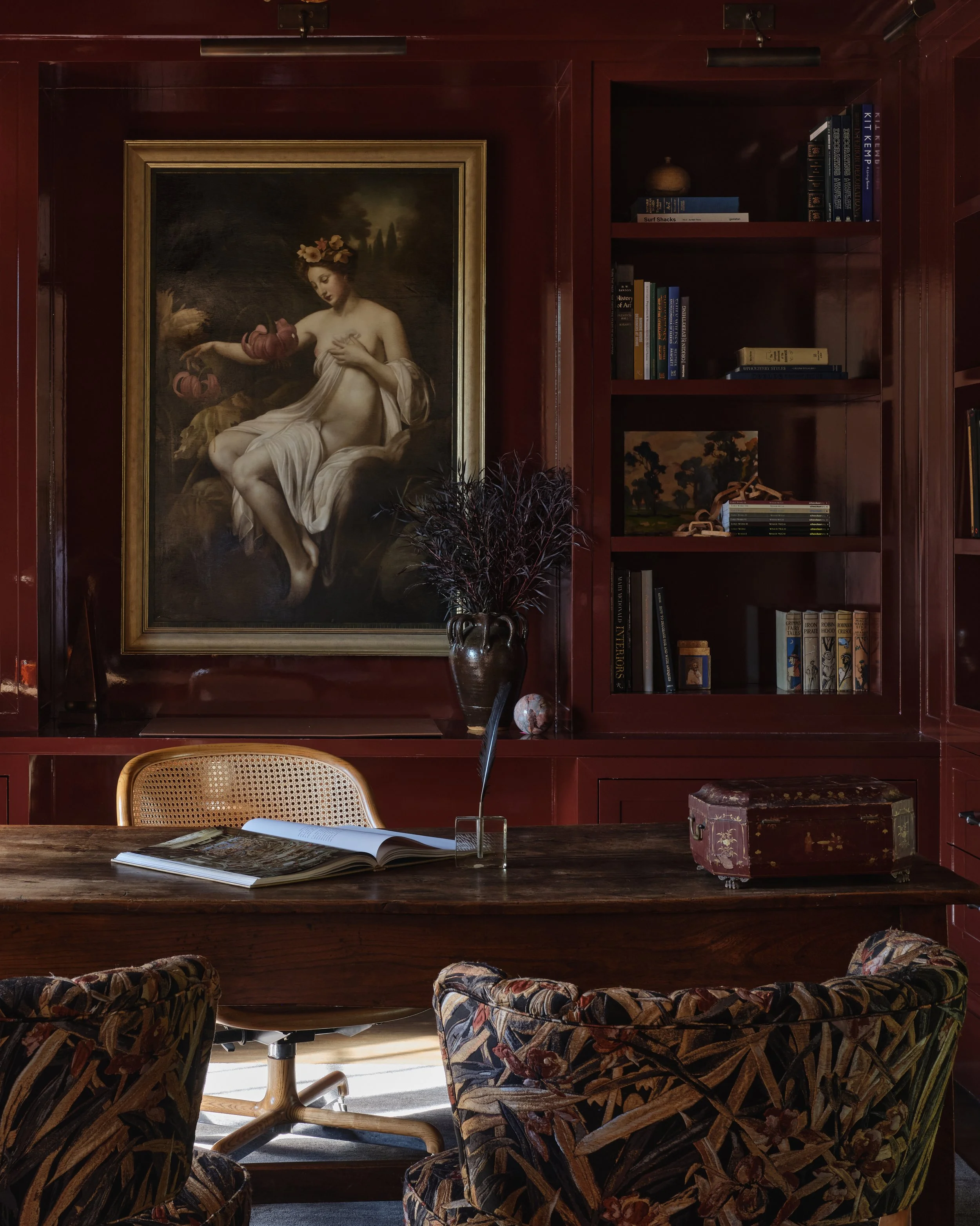 A room with dark red wood walls and shelves, a large framed classical painting of a nude woman with a floral crown, a wooden desk with an open book, a glass vase with dark foliage, vintage chairs, and a decorative box.