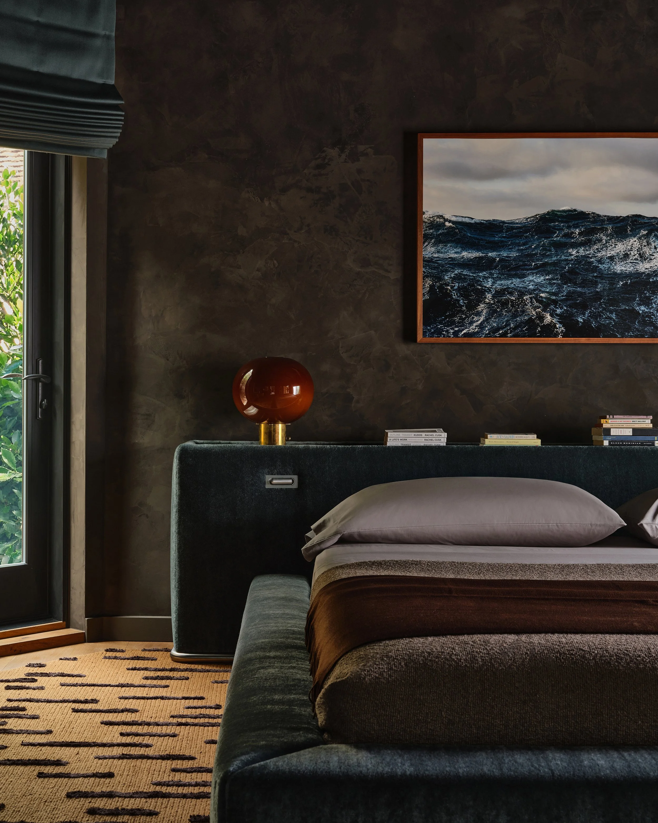 Comedian Anthony Jeselnik’s bedroom room, interior design by Riccio Vidaurre Design – A dark and moody bedroom with brown plaster walls and custom a blue mohair platform bed.  An orange lamp sits on the headboard with a photograph of the ocean.
