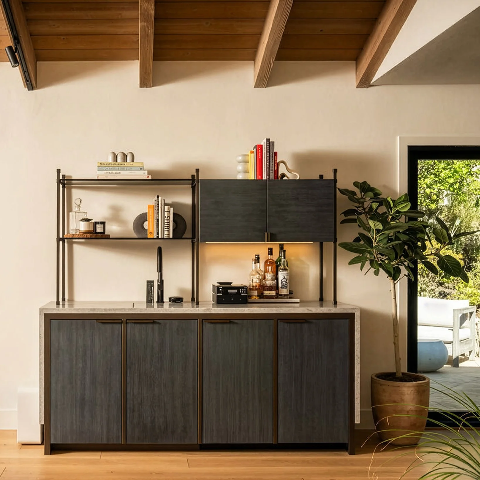 A wet bar with blue-grey stained wood, brass frame and stone countertop.  Glass and brass shelves sit ontop, styled with alcohol bottles and books.