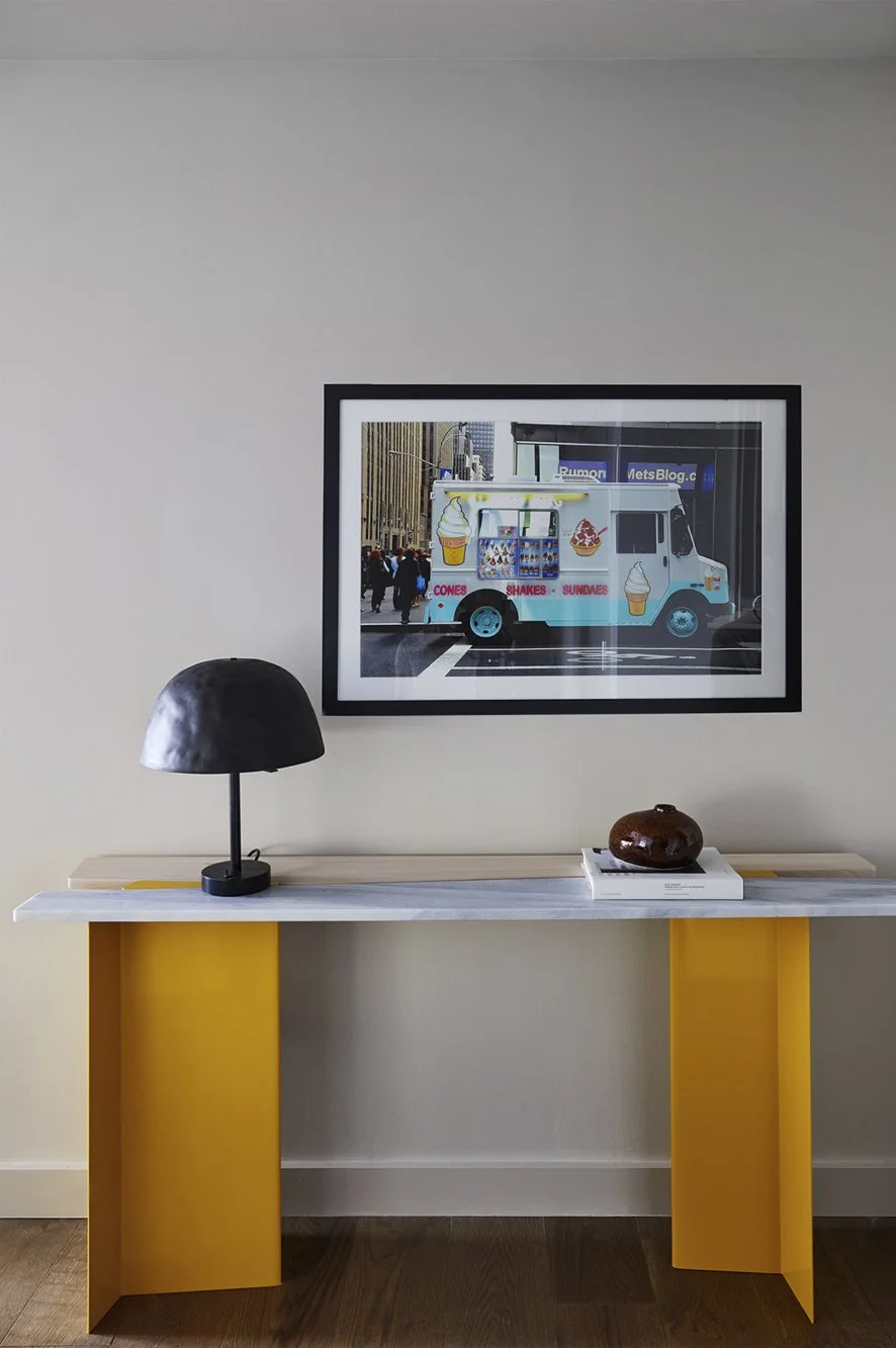 Decorative wall art of an ice cream truck in an interior setting on a white wall, with a modern yellow and white console table below it. The table has a black desk lamp and a small brown vase on top.