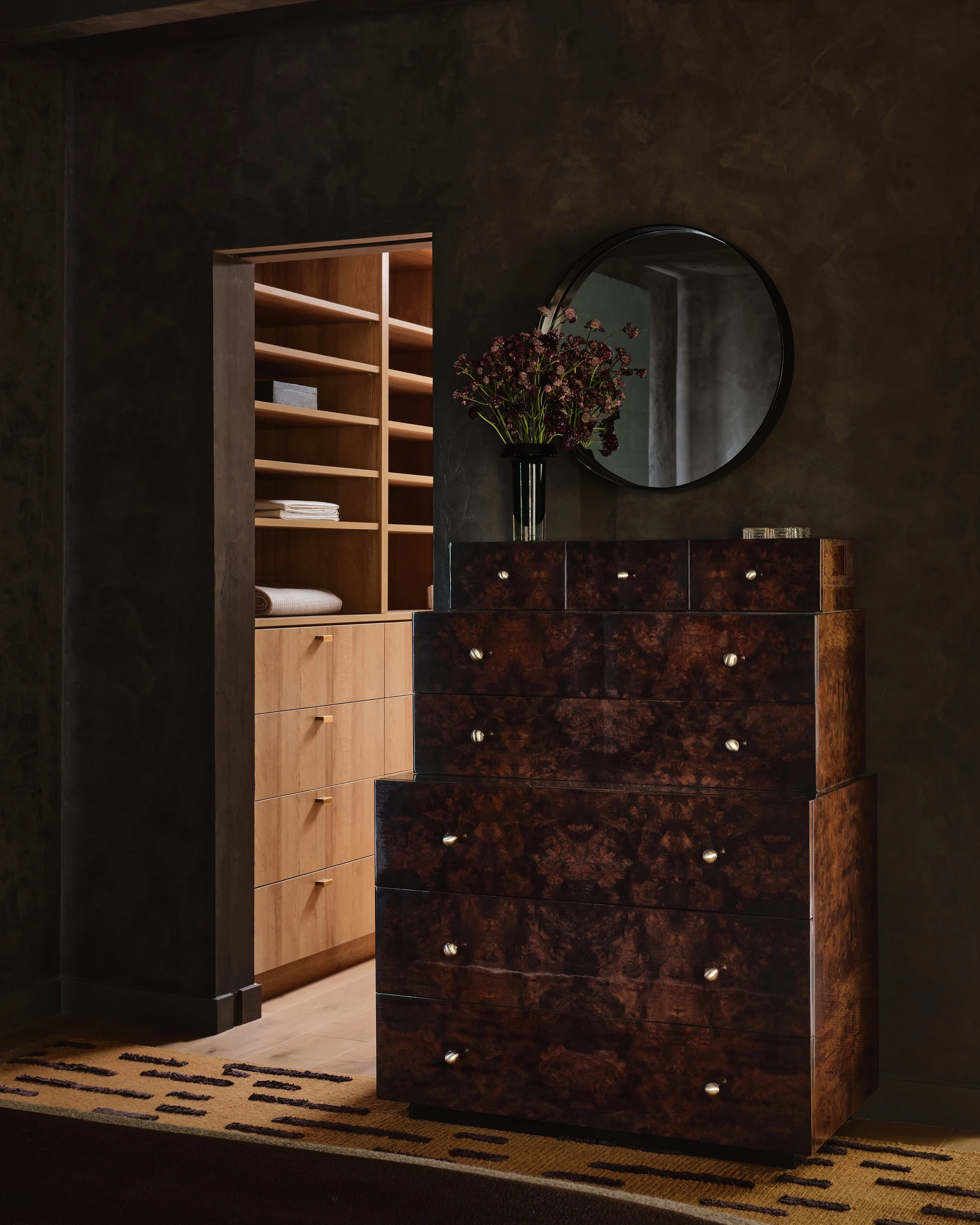Comedian Anthony Jeselnik’s bedroom room, interior design by Riccio Vidaurre Design – A dark and moody bedroom with brown plaster walls and a burl wood dresser that steps back with a metal circle mirror above it. A doorway to a closet.