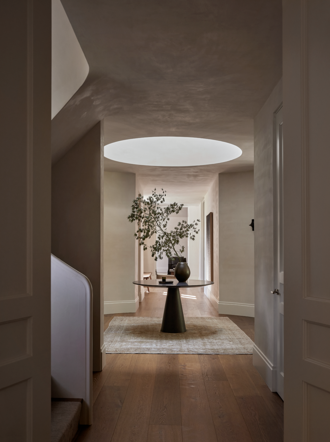 Interior view of a modern hallway with a round skylight, a round table with a vase and plant, and wooden flooring.
