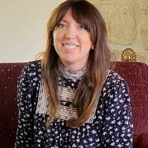 A woman with brown hair and a smile, wearing a patterned sweater, sitting on a red upholstered chair in a room with stone walls and decorative items.