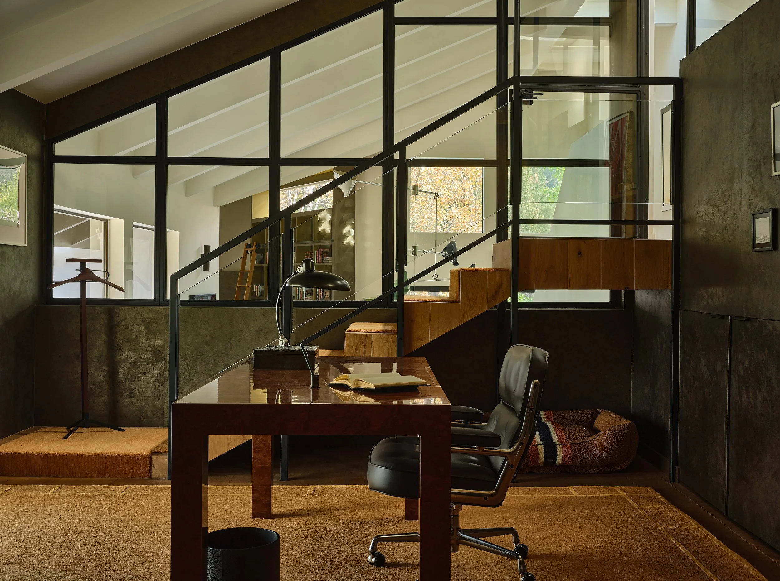 Anthony Jeselnik’s home office, interior design by Riccio Vidaurre Design – the walls are a bronze wallpaper and their are interior windows.  There is a large burl parsons desk with a black desk chair and orange carpet.