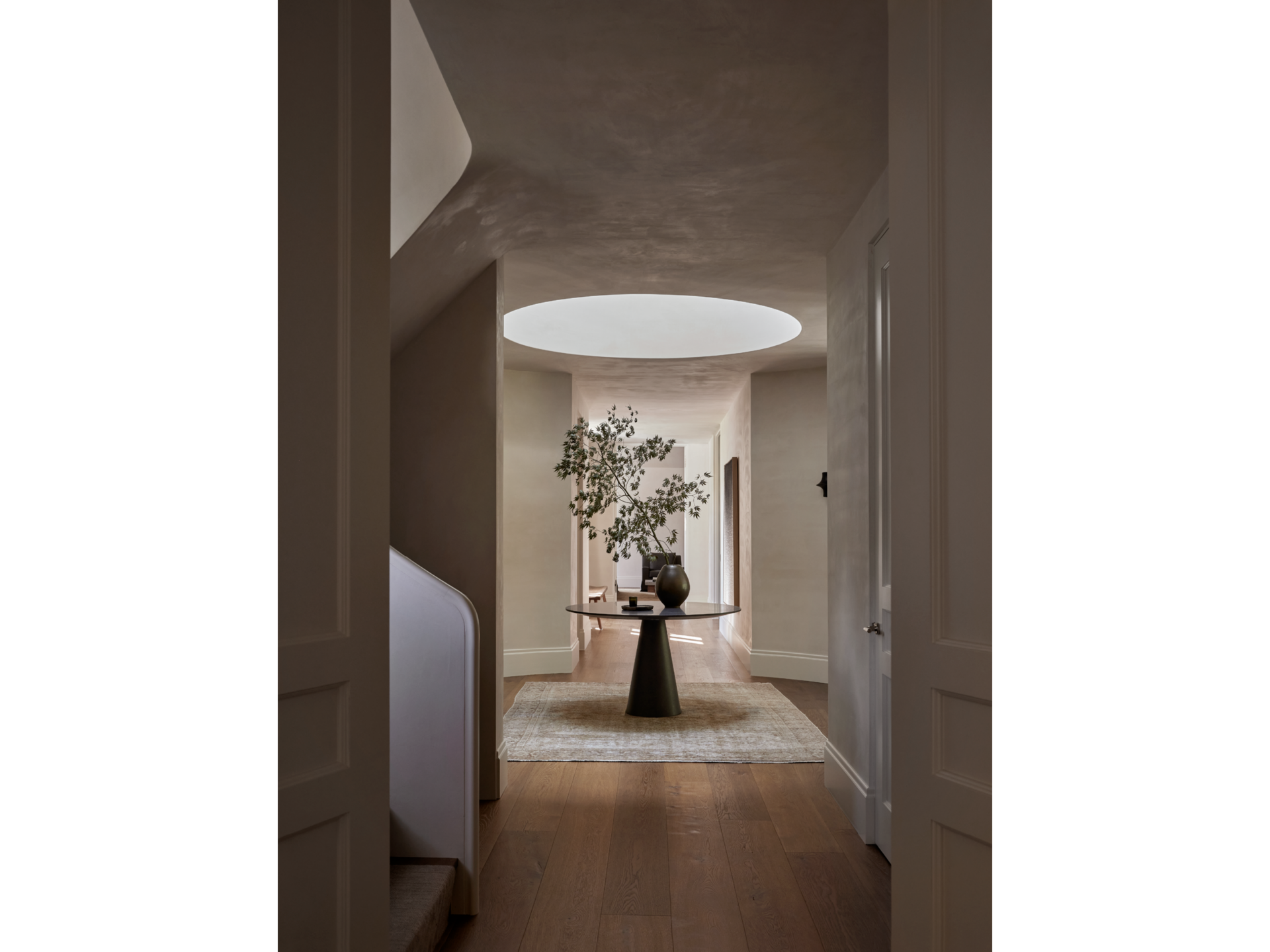 Minimalist interior hallway leading to a round table with a vase and a tree inside, illuminated by a circular skylight overhead.