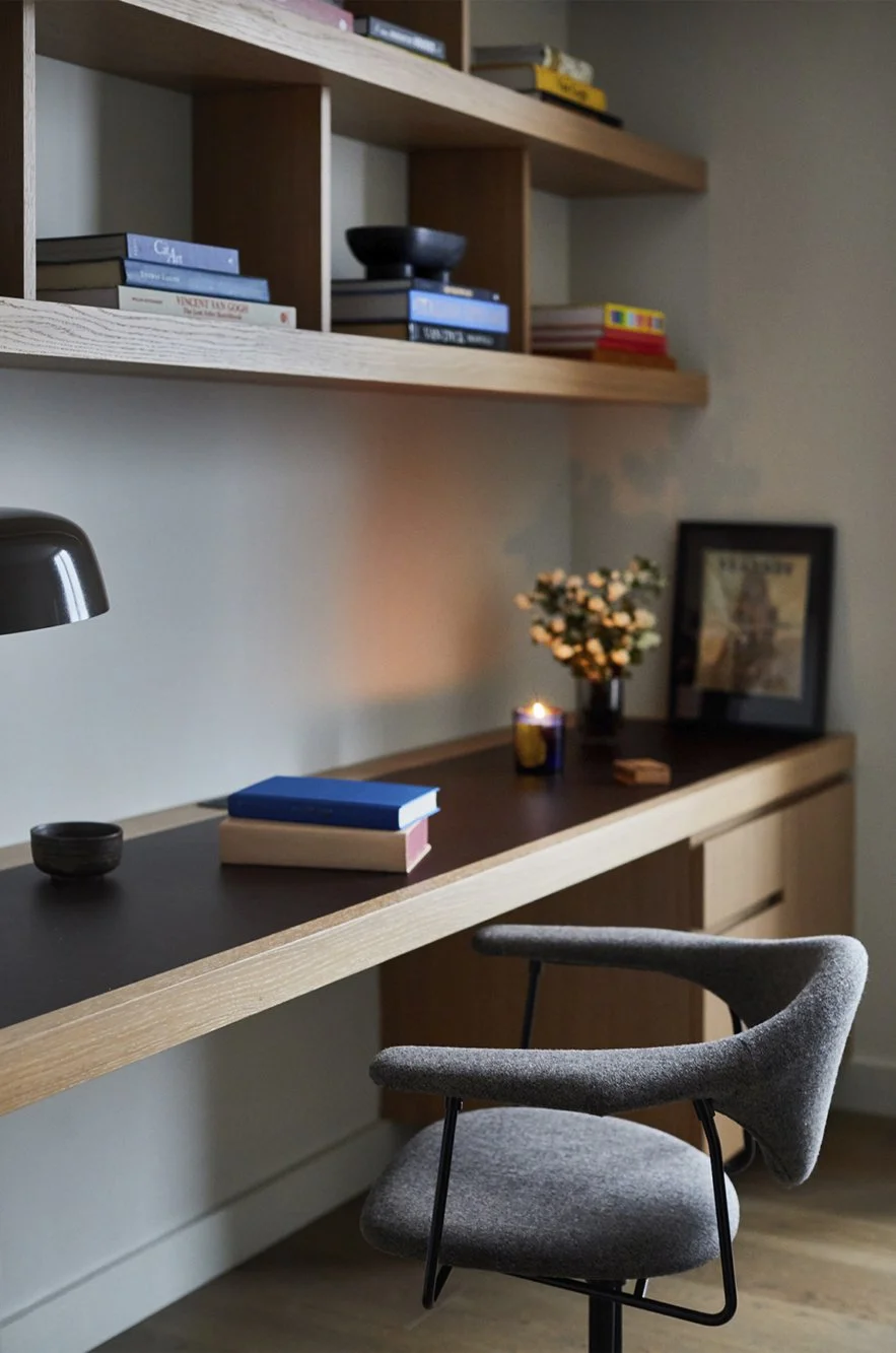 A modern workspace with a wooden desk, books, a plant, a candle, framed art, and a gray upholstered chair.