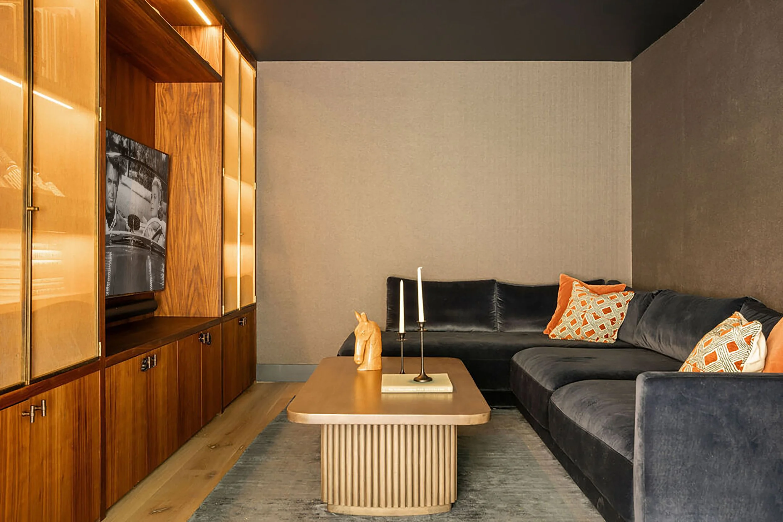 A moody den with grey wallpaper, dark grey ceiling and walnut and brass built-ins that house the television.  There is a blue velvet sectional sofa with a wood coffee table styled with candlesticks, books and a horse statue.