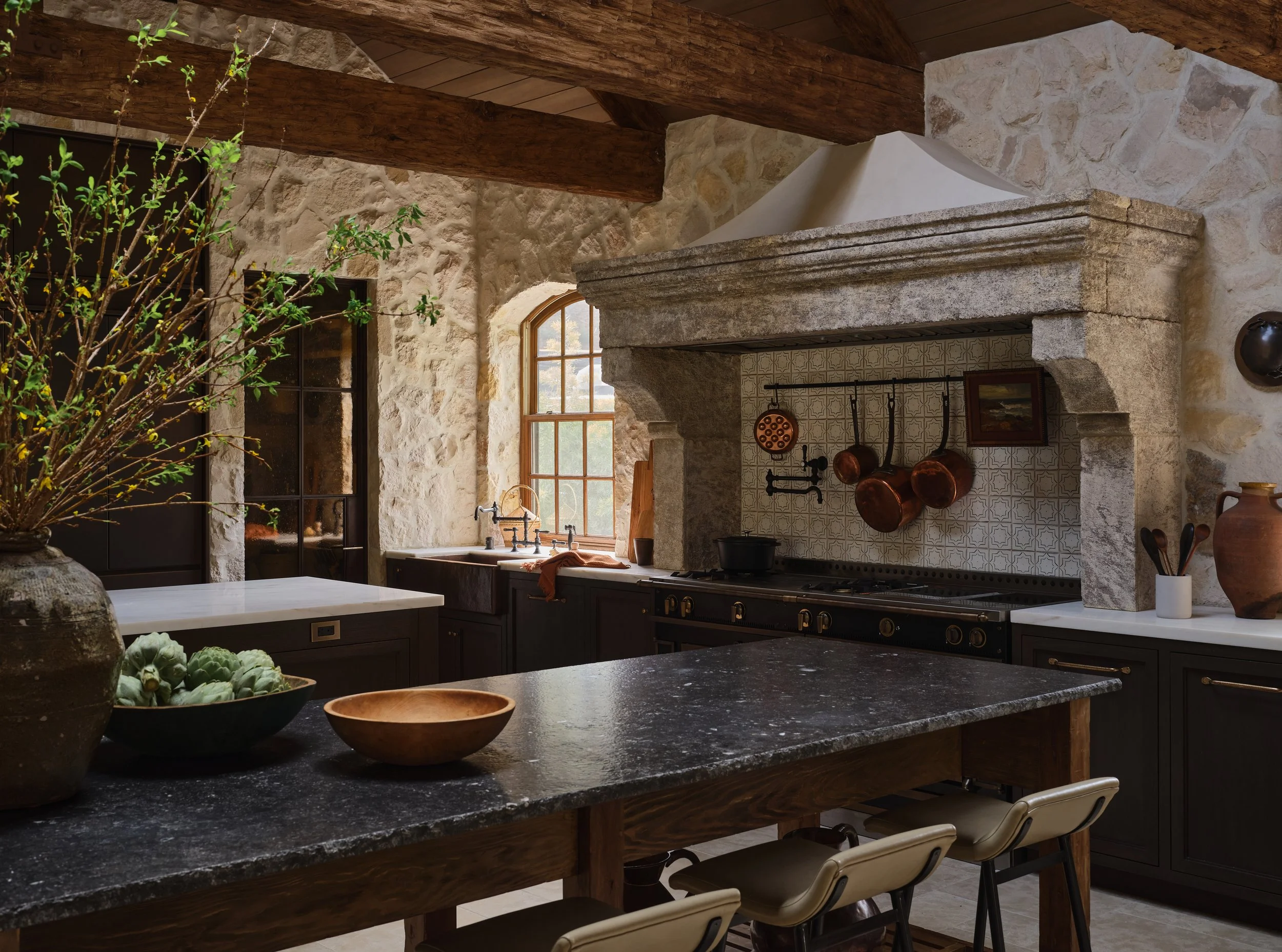 Rustic kitchen with stone walls, wooden beams, black cabinetry, a large granite island, a vintage stove with hanging pots, and a window with a scenic view.