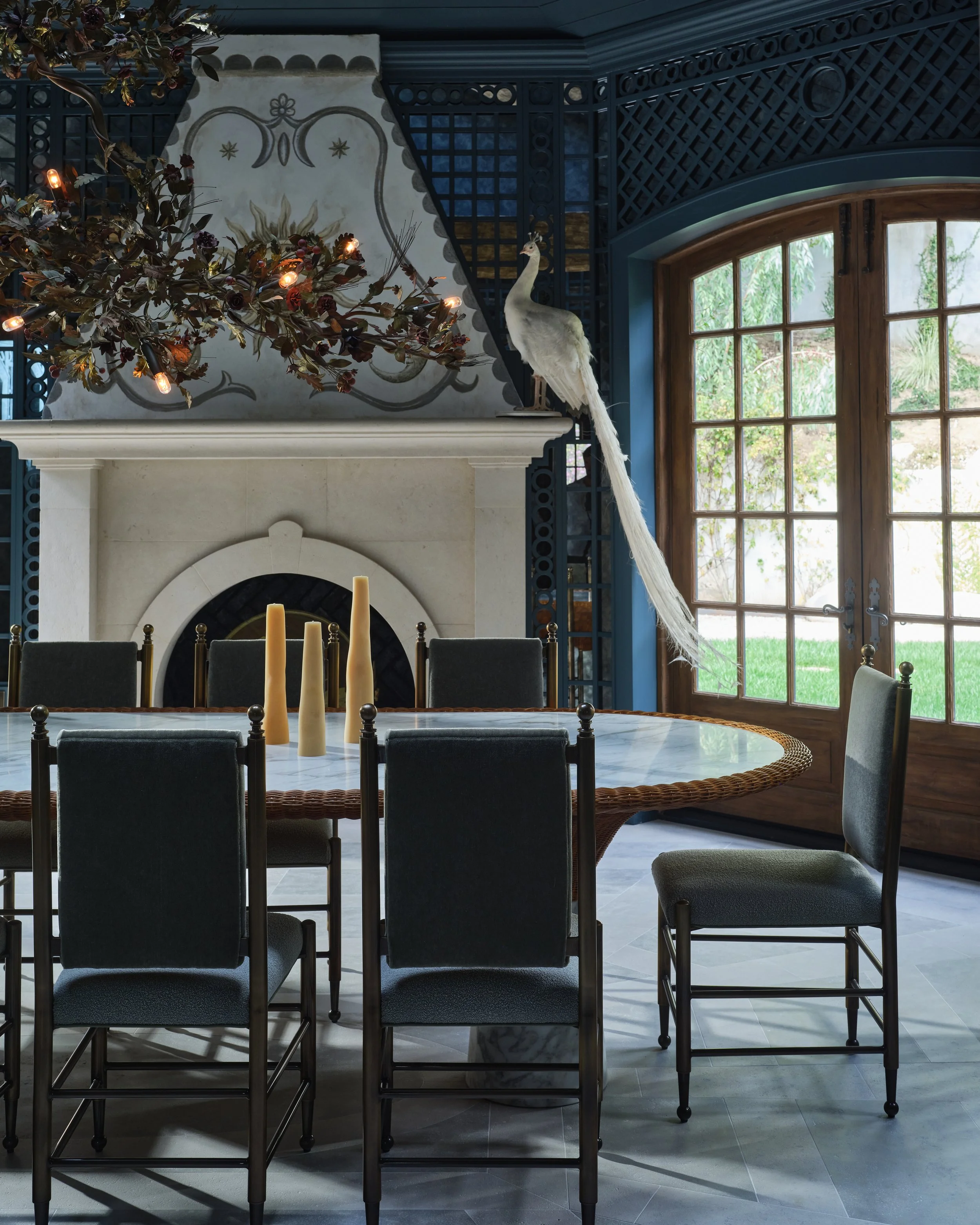 A dining room with a round marble table, six dark upholstered chairs, three beige candles in vases, a decorative white fireplace, and a peacock perched on the mantle near large windows with wooden frames and glass panes, overlooking a garden.