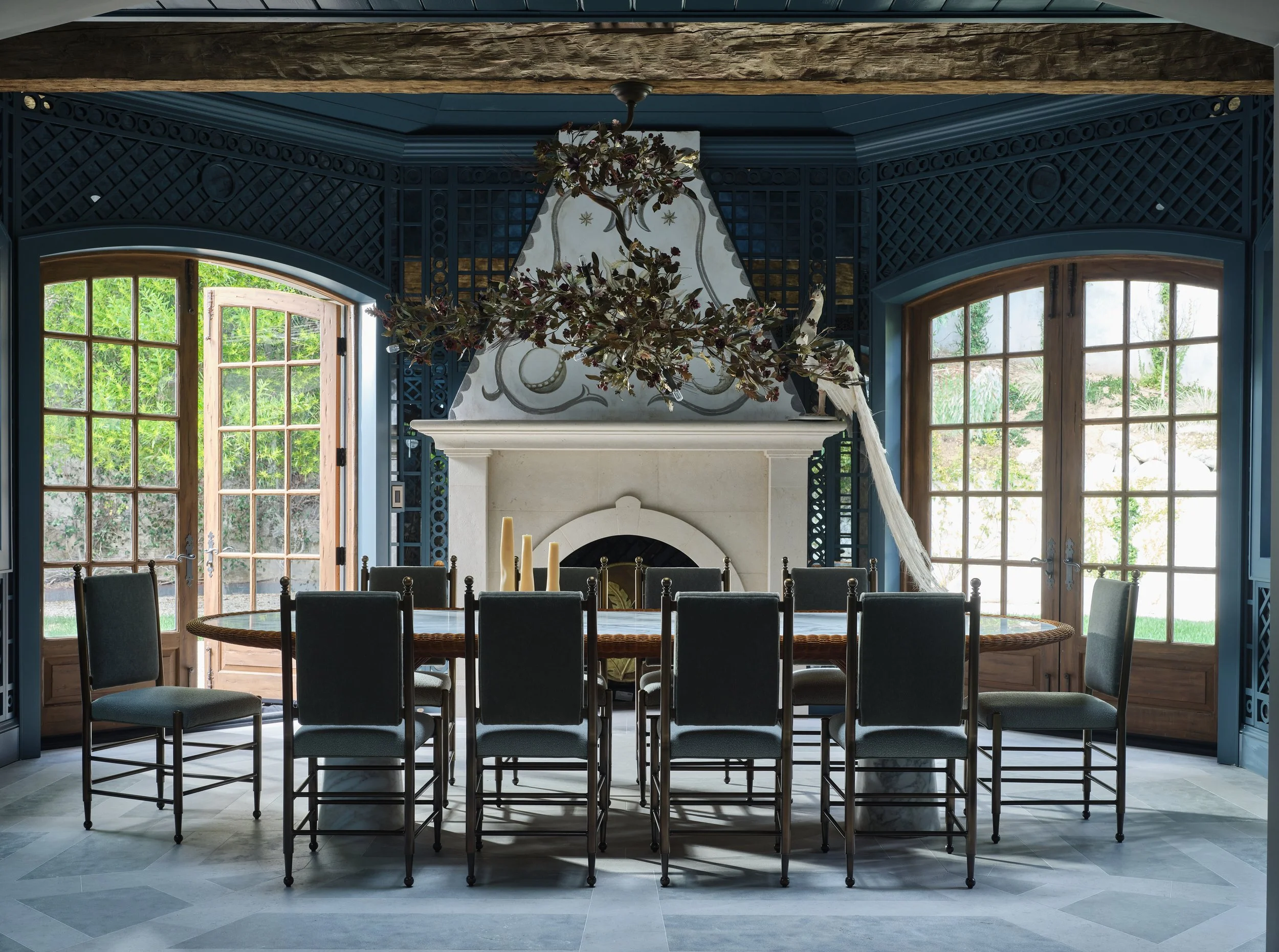Dining room with a large round table, ten chairs, a fireplace, and large windows with wooden frames, decorated with a floral chandelier.