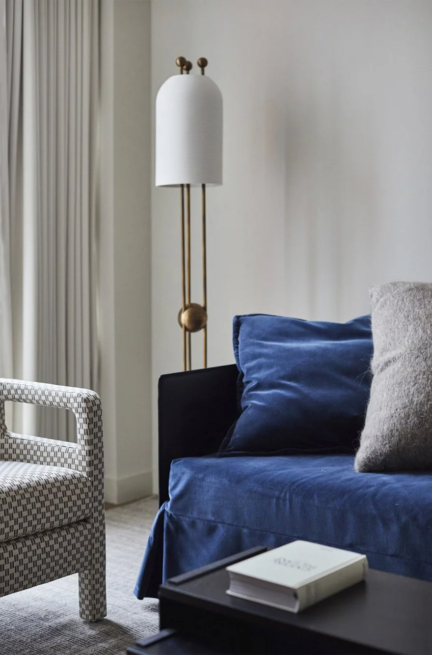 Living room corner with a patterned armchair, a navy sofa with cushions, a side table with a book, a tall white and brass floor lamp, and curtains along the wall.