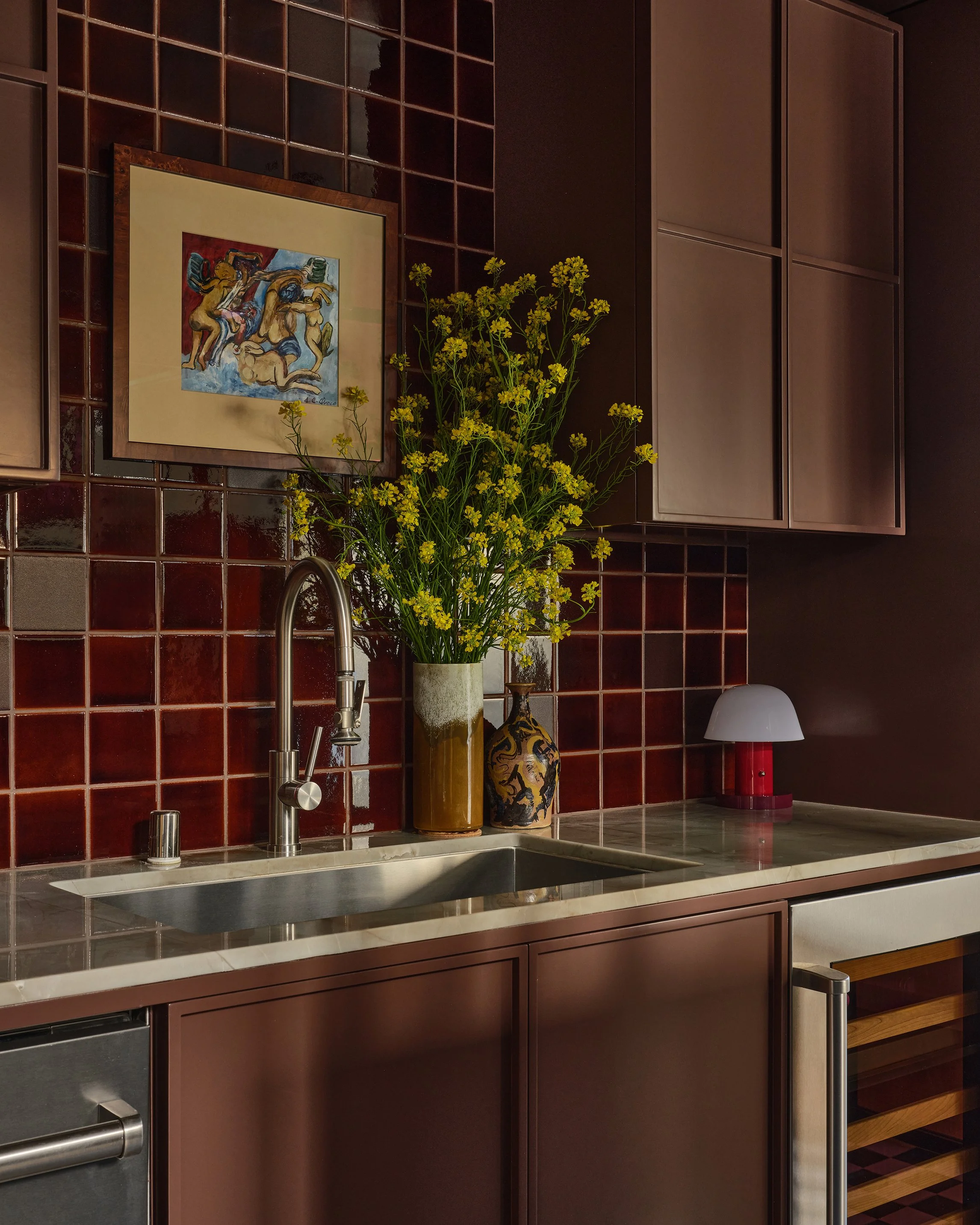 Comedian Anthony Jeselnik’s home by Riccio Vidaurre Design – A kitchen sink with brown painted cabinets and brown square ceramic tiles backsplash.  A painting hangs above the sink, there is a vase of yellow wildflowers and a small red lamp.