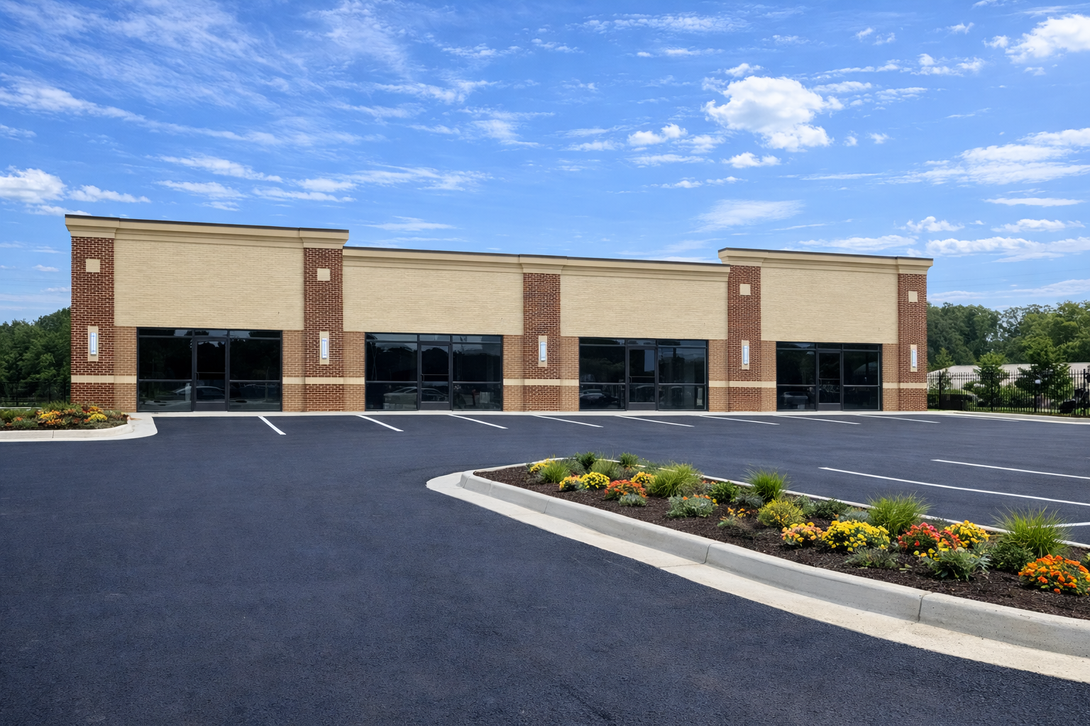 Newly constructed single-story commercial building with glass storefronts, a black paved parking lot with white lines, and landscaped islands with colorful flowers and green plants under a blue sky with scattered clouds.