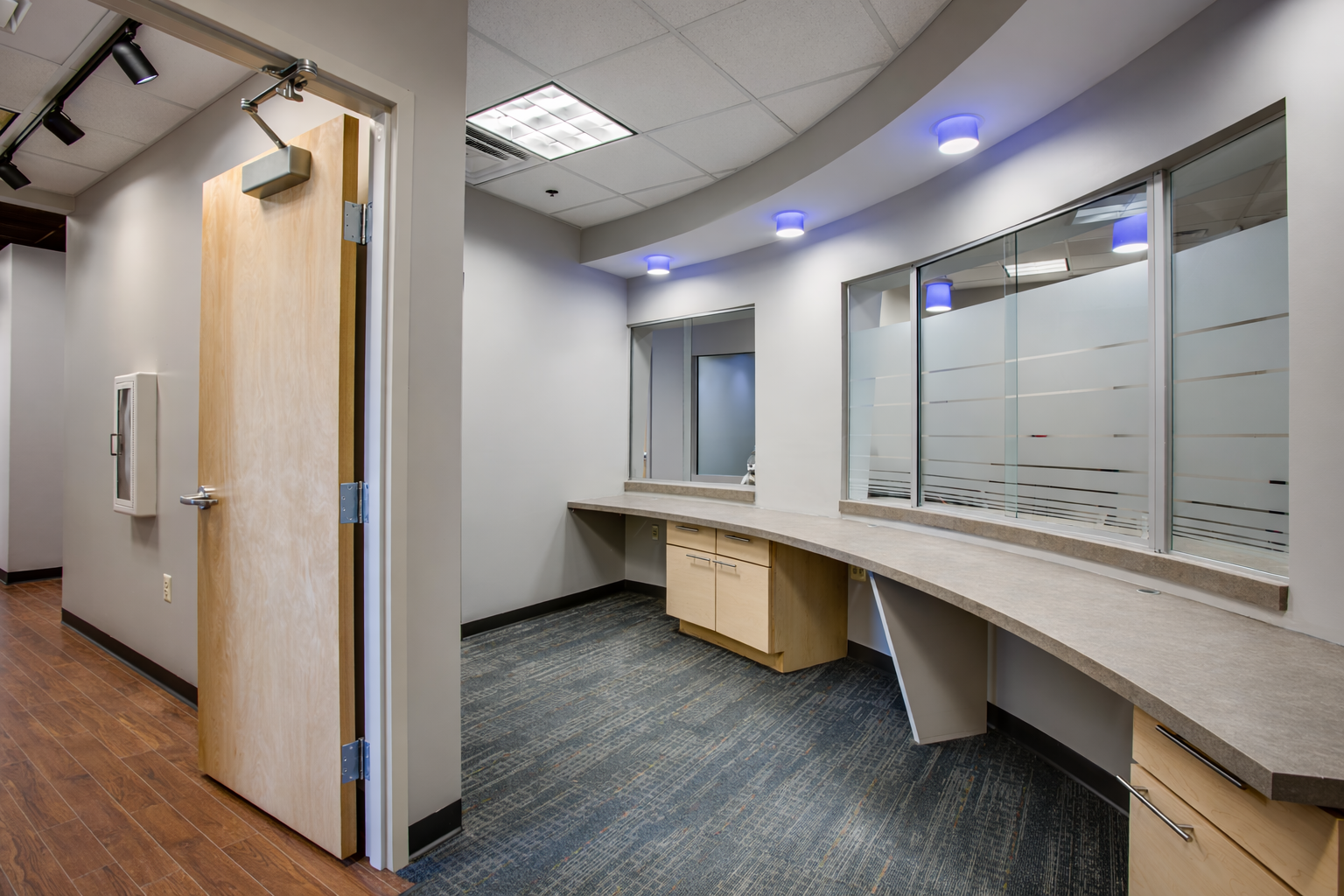 Empty office reception area with a curved counter, windowed partitions, overhead blue and white lighting, wood flooring, and a wooden door.