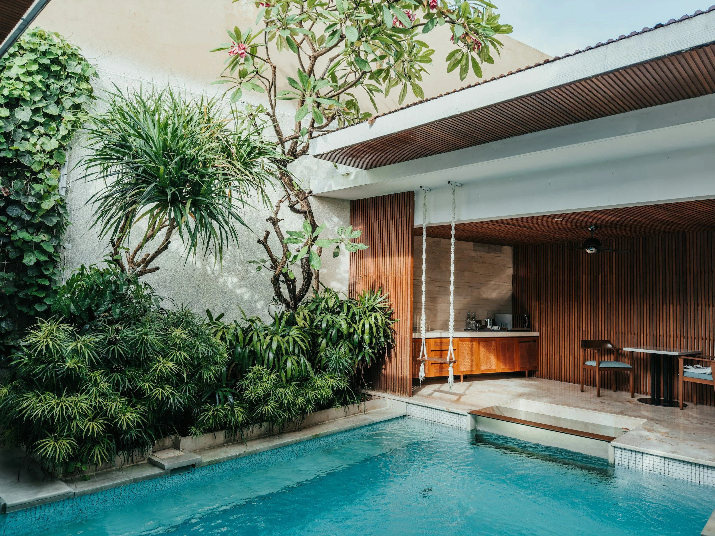 An outdoor swimming pool surrounded by lush green plants and trees, with a modern open-air bar area featuring wood paneling and a ceiling fan.