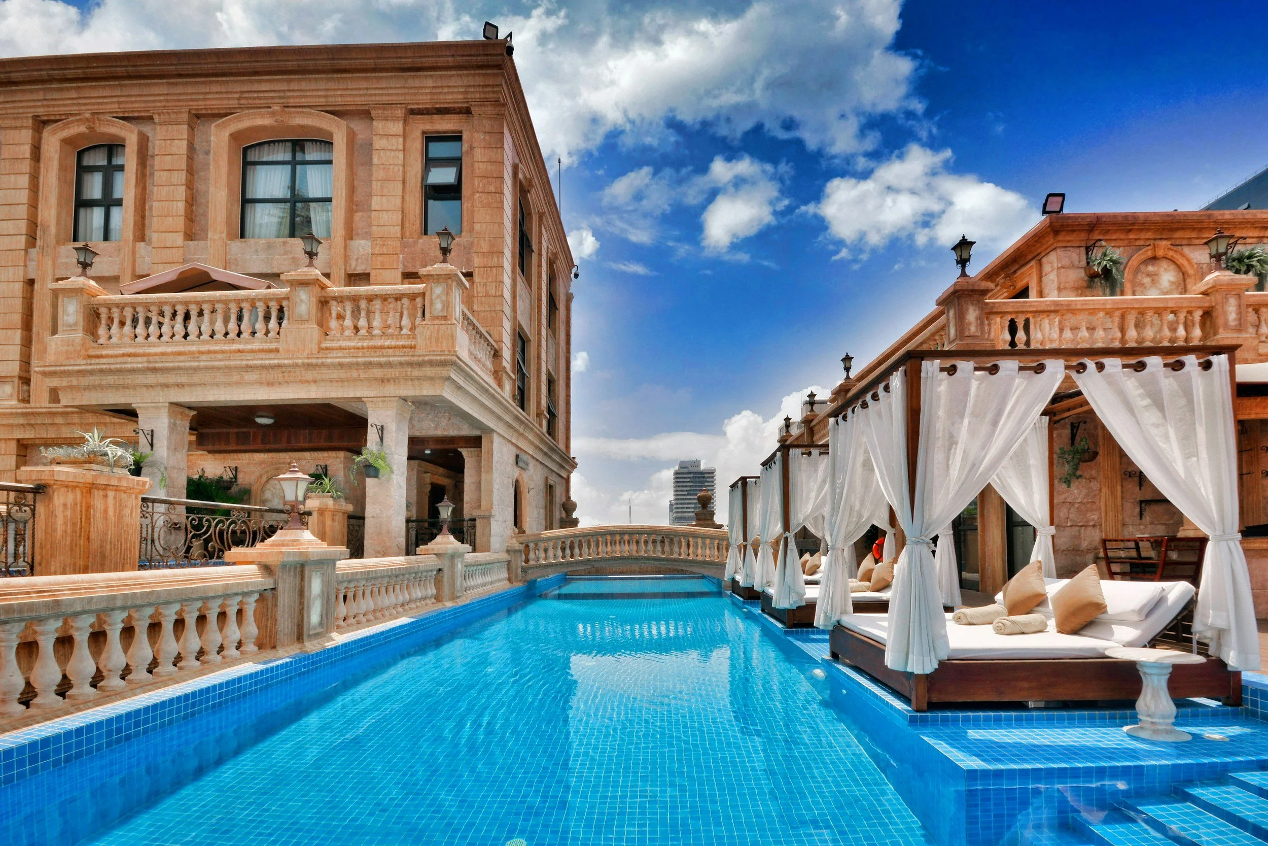 Upscale rooftop scene with a swimming pool, white cabanas with curtains, stone building with balconies and potted plants, blue sky with clouds.