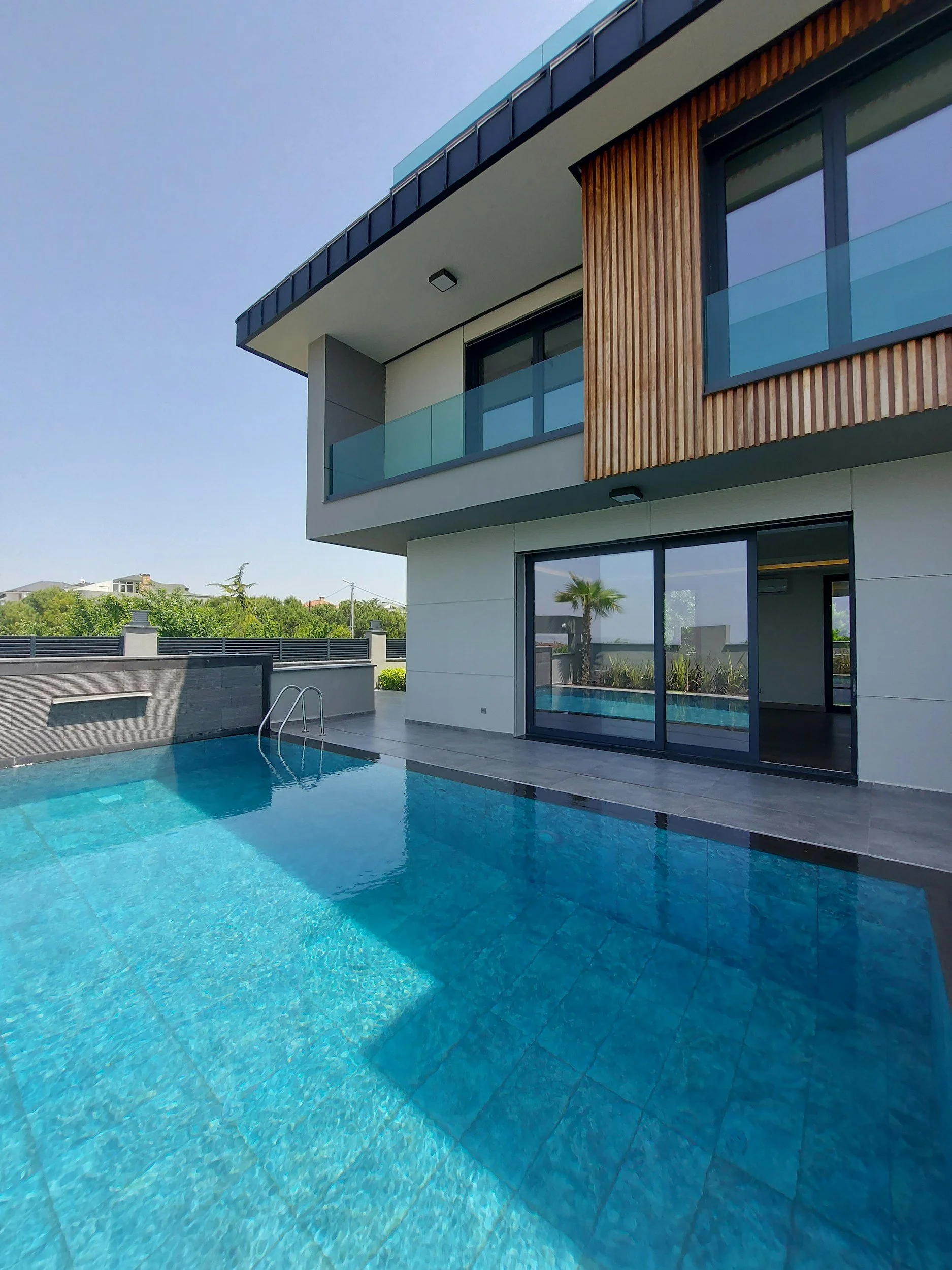Modern house with a swimming pool, glass balcony, and wooden exterior panels, under a clear blue sky.
