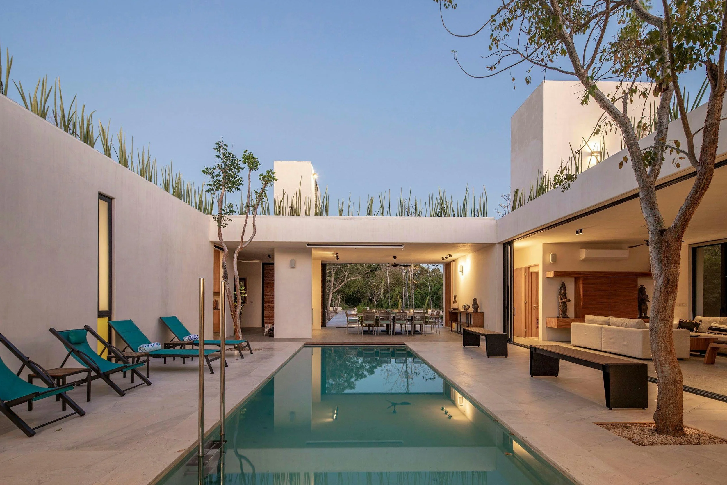 Modern house patio with a swimming pool, lounge chairs, and an outdoor dining area, surrounded by trees and minimalist white walls, during evening with warm indoor lighting.