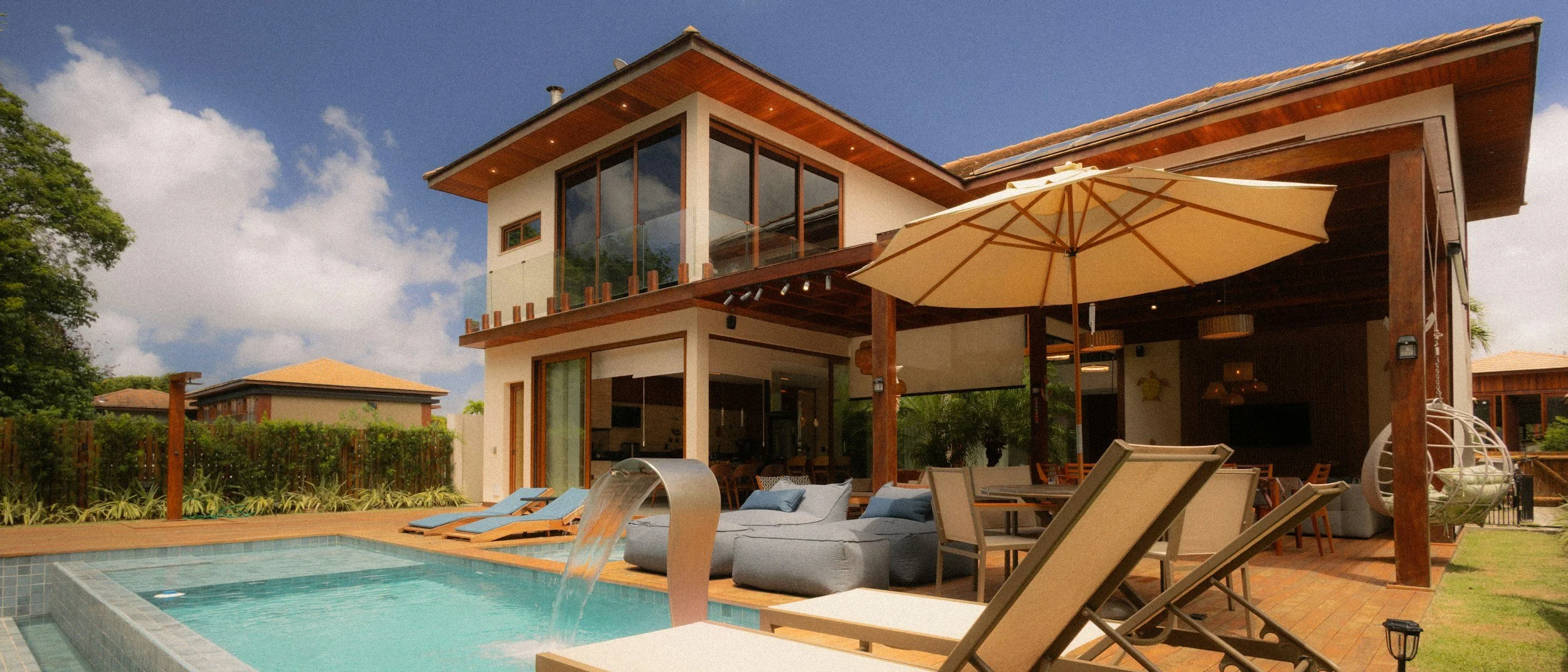Modern two-story house with a pool and outdoor seating area, including lounge chairs, a large umbrella, and a hanging chair, under a partly cloudy sky.