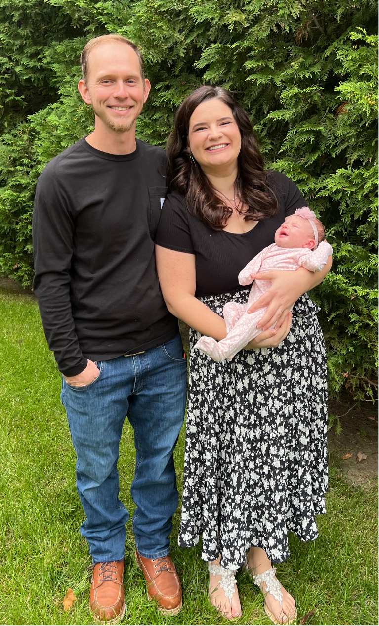 Devin Bilello and Jackie Bilello holding a baby girl outdoors in front of green shrubbery.
