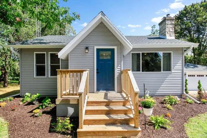 A modern house with light gray siding, a blue front door, and a gable roof. Wooden stairs lead to the front porch, surrounded by landscaped garden beds with flowering plants and mulch. DRB Services home remodel hvac systems.