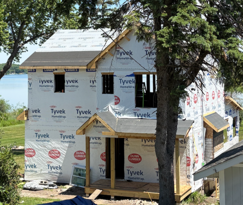 A house under construction wrapped in Tyvek HomeWrap, with a large tree in front, near a body of water. DRB Services new construction hvac systems.
