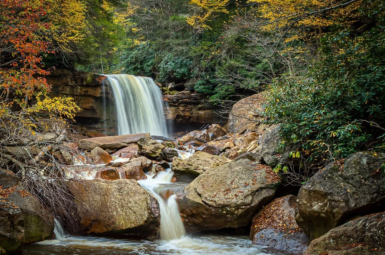 A small waterfall flowing over rocks in a forest with trees showing fall foliage.