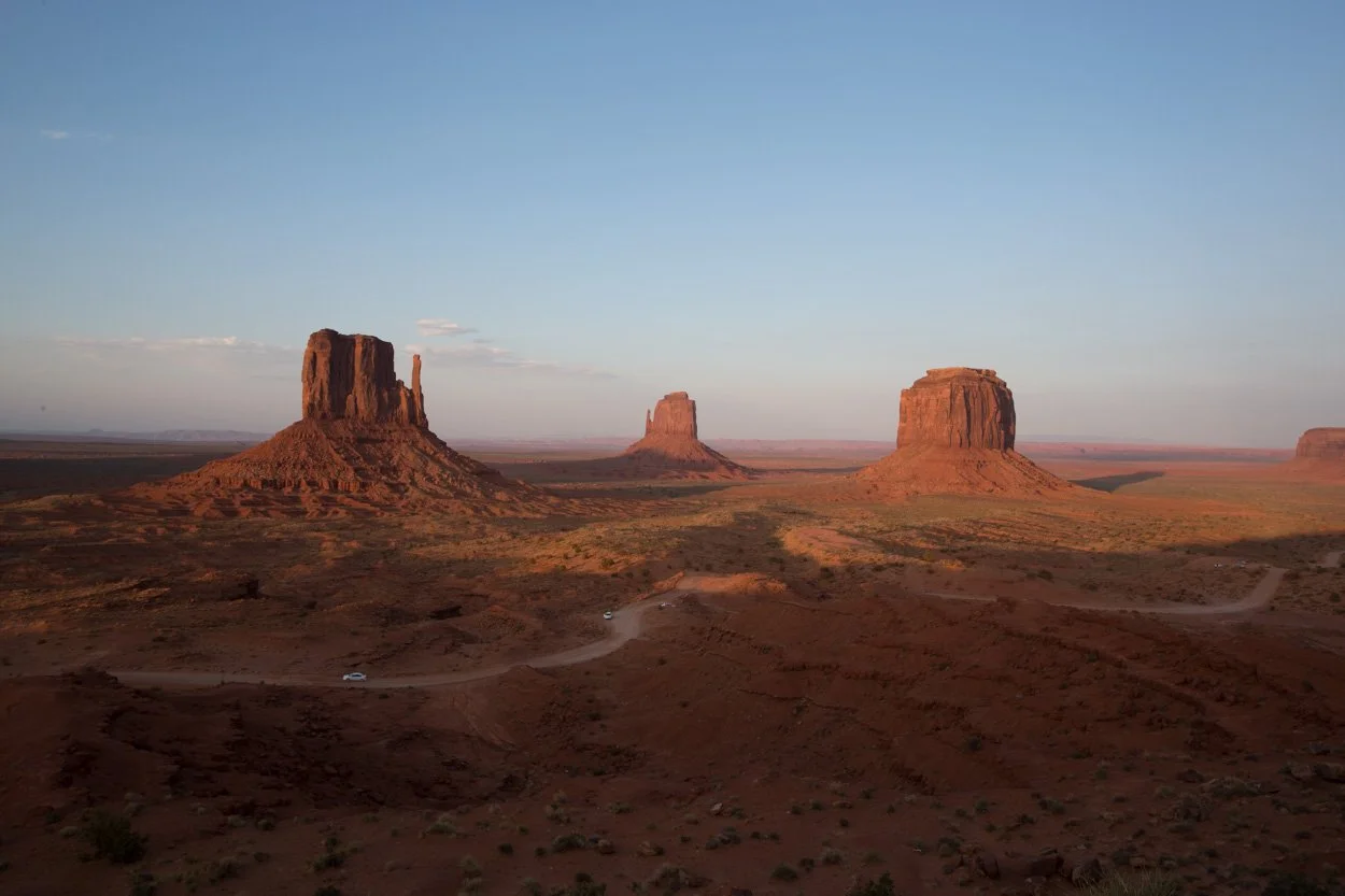 Three large desert rock formations known as buttes, with a dirt road winding through a red landscape, under a clear blue sky, in Monument Valley.