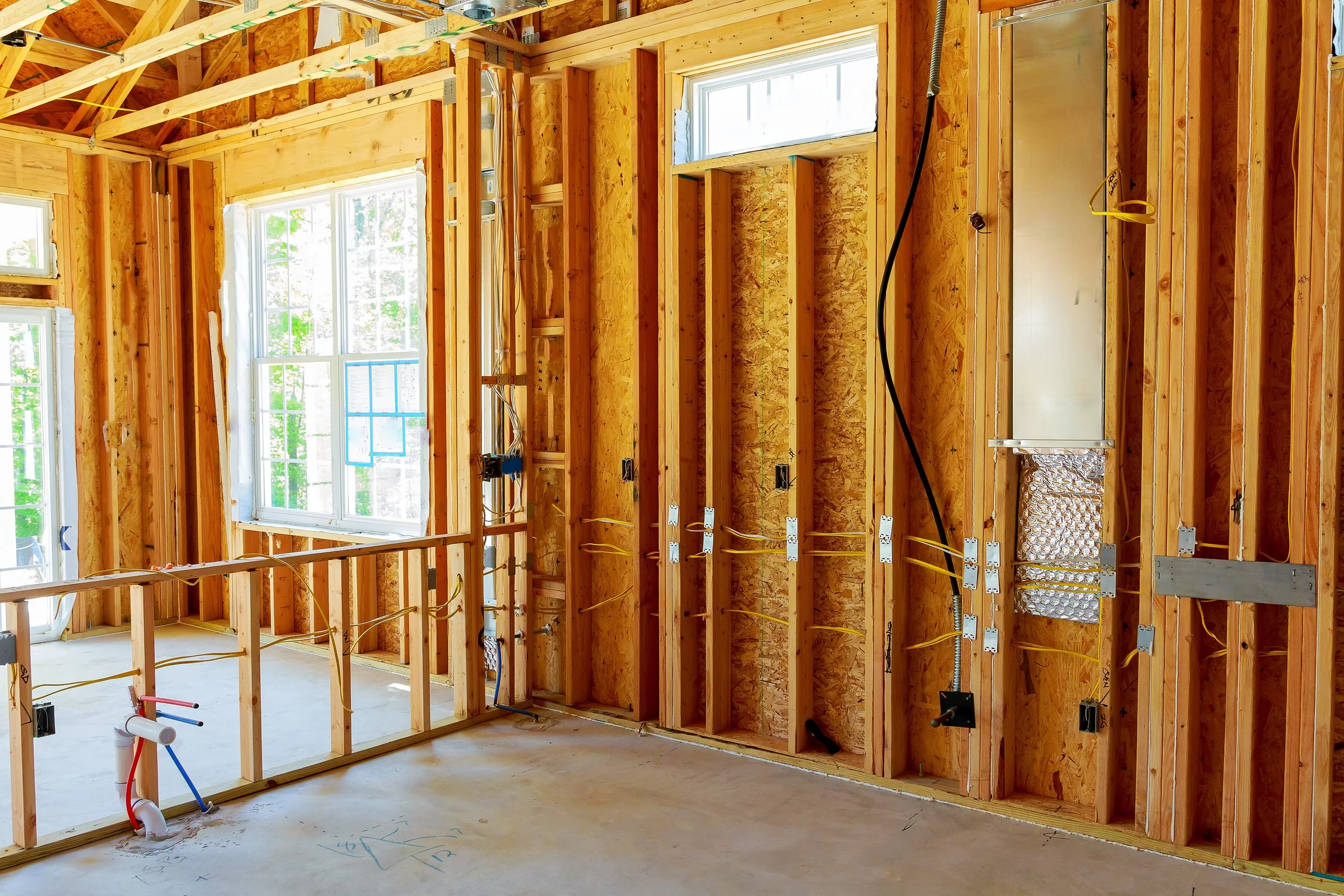 Interior view of a house under construction, showing exposed wooden framing, electrical wiring, and windows.