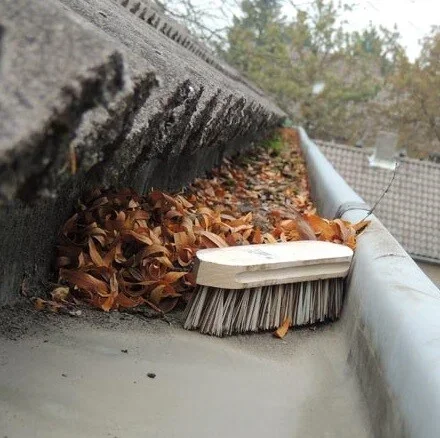 Een dakgoot met bladeren en een borstel ernaast, klaar om gereinigd te worden. 