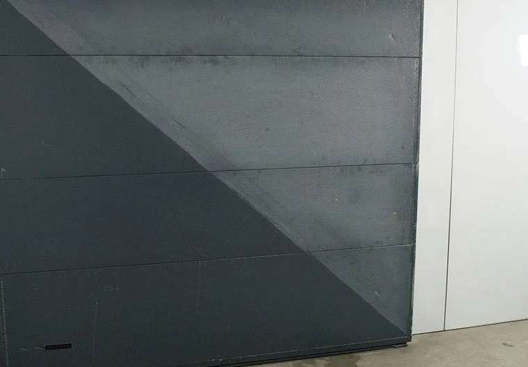 Close-up van een garagepoort met diagonale kleurverschillen in grijstinten. De ene helft heeft een reiniging gehad. De andere helft moet nog gereinigd worden. 