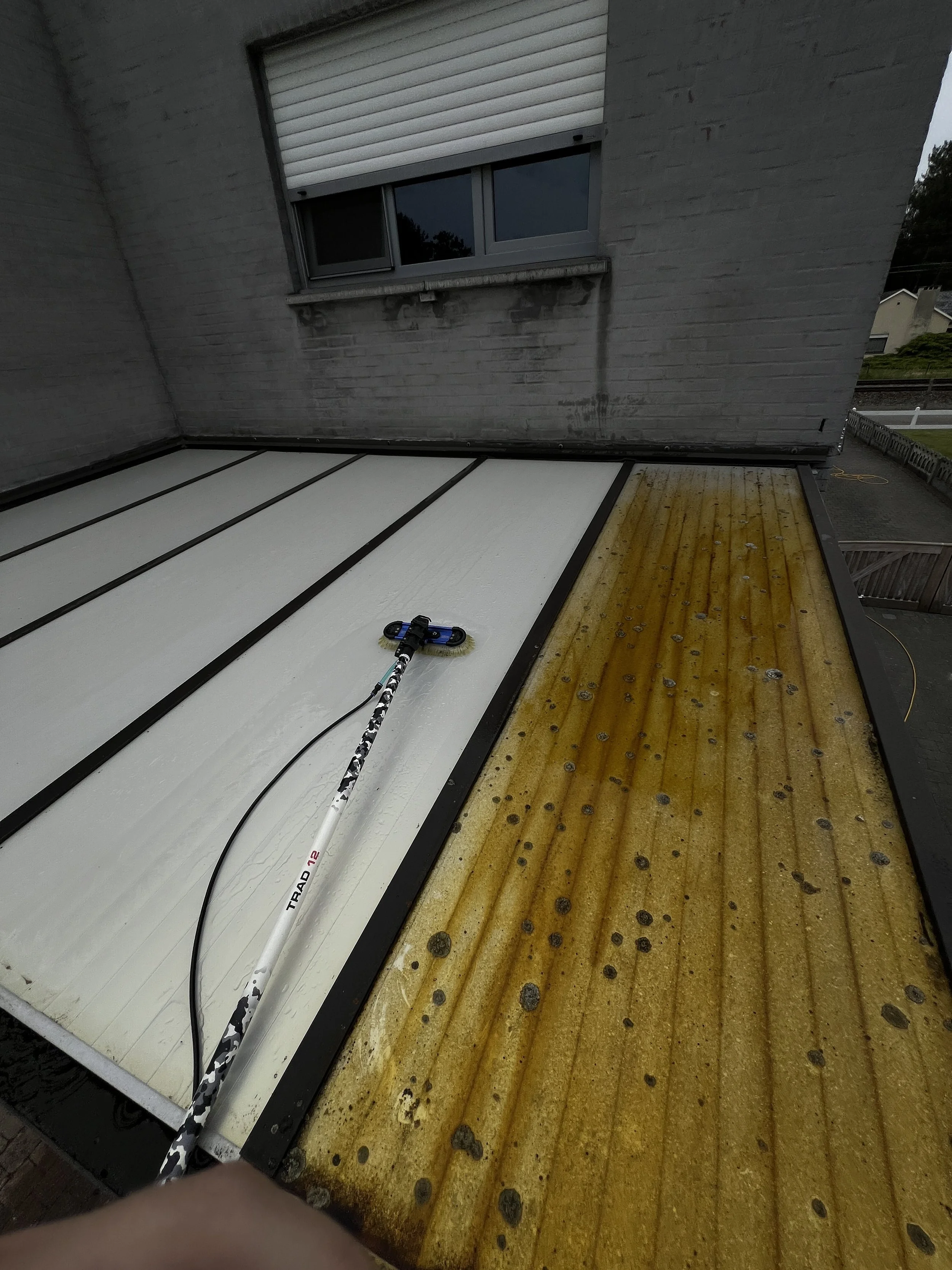 Op het platte afdak worden schoonmaakwerkzaamheden uitgevoerd. Het dak is deels wit na reiniging en deels geel/bruin door vervuiling en schimmel.