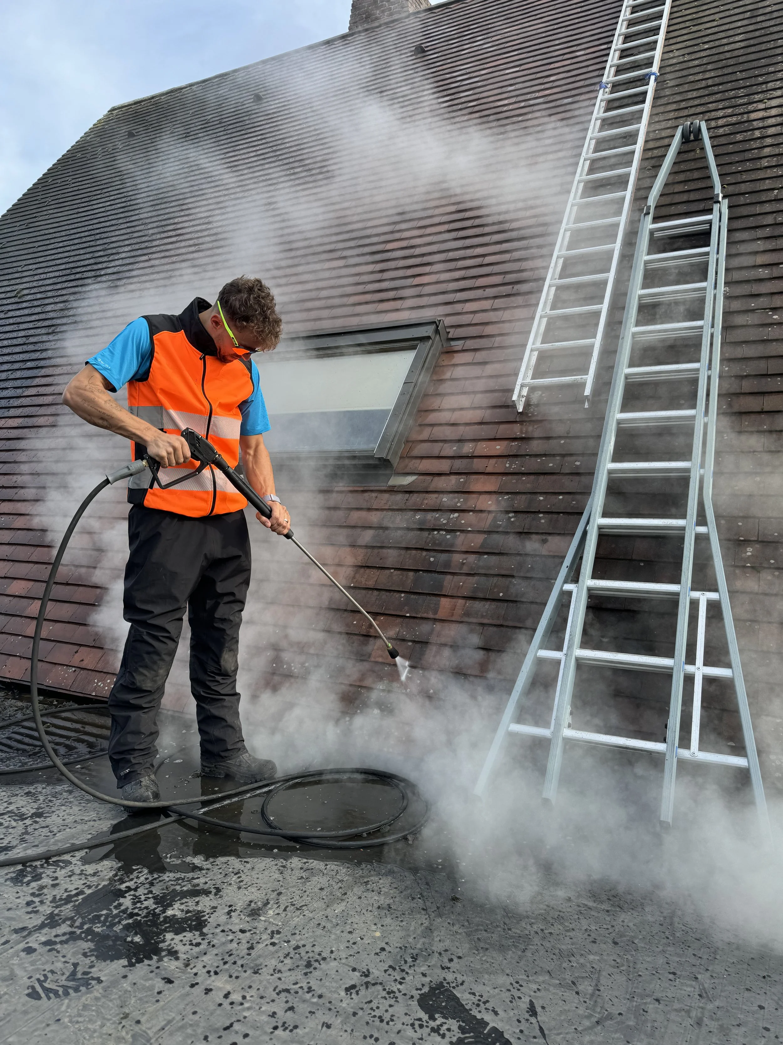 Man gebruikt een stoomreiniger op een pannendak, met wolk van waterdamp en een ladder op het dak.