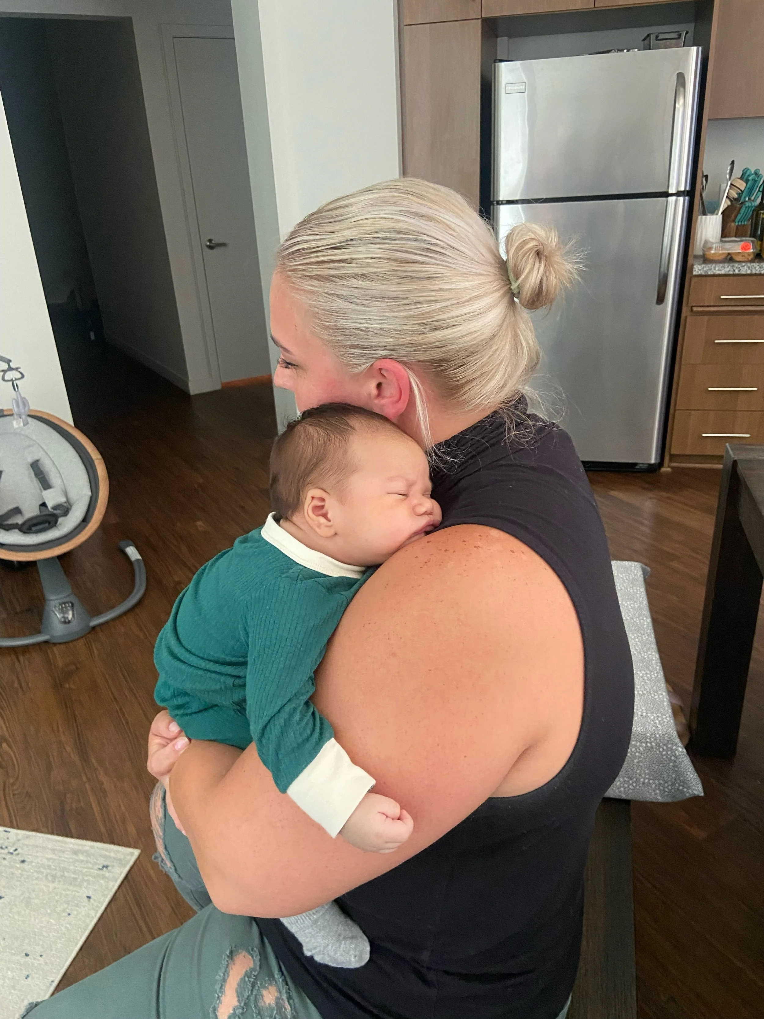 A woman with blonde hair tied back, holding a sleeping baby with brown hair, in a kitchen with wooden cabinets and a silver refrigerator.
