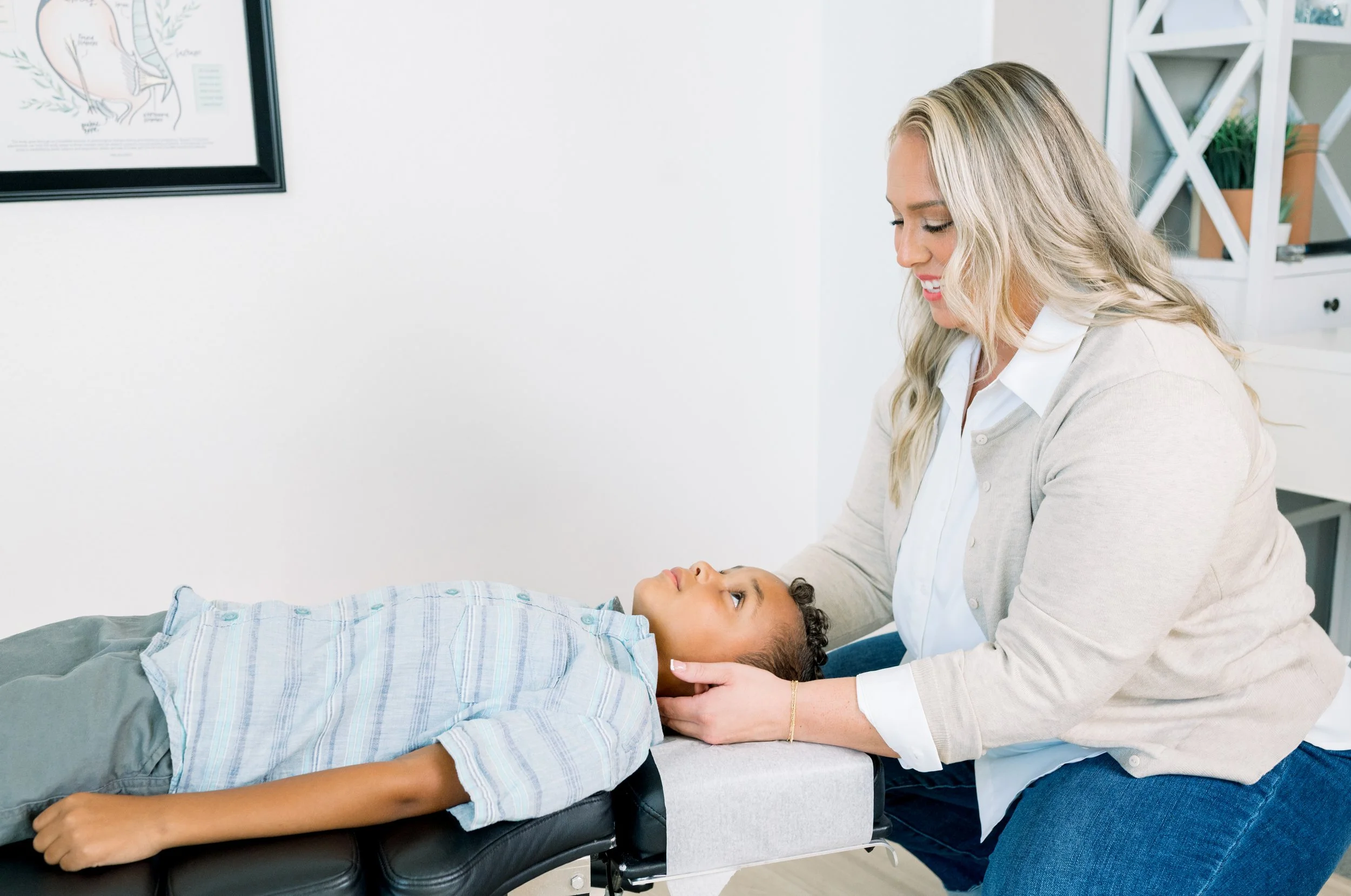A chiropractor with blonde hair, wearing a white shirt and beige cardigan, gently checking a child's neck during a consultation or examination in a chiropractic office.